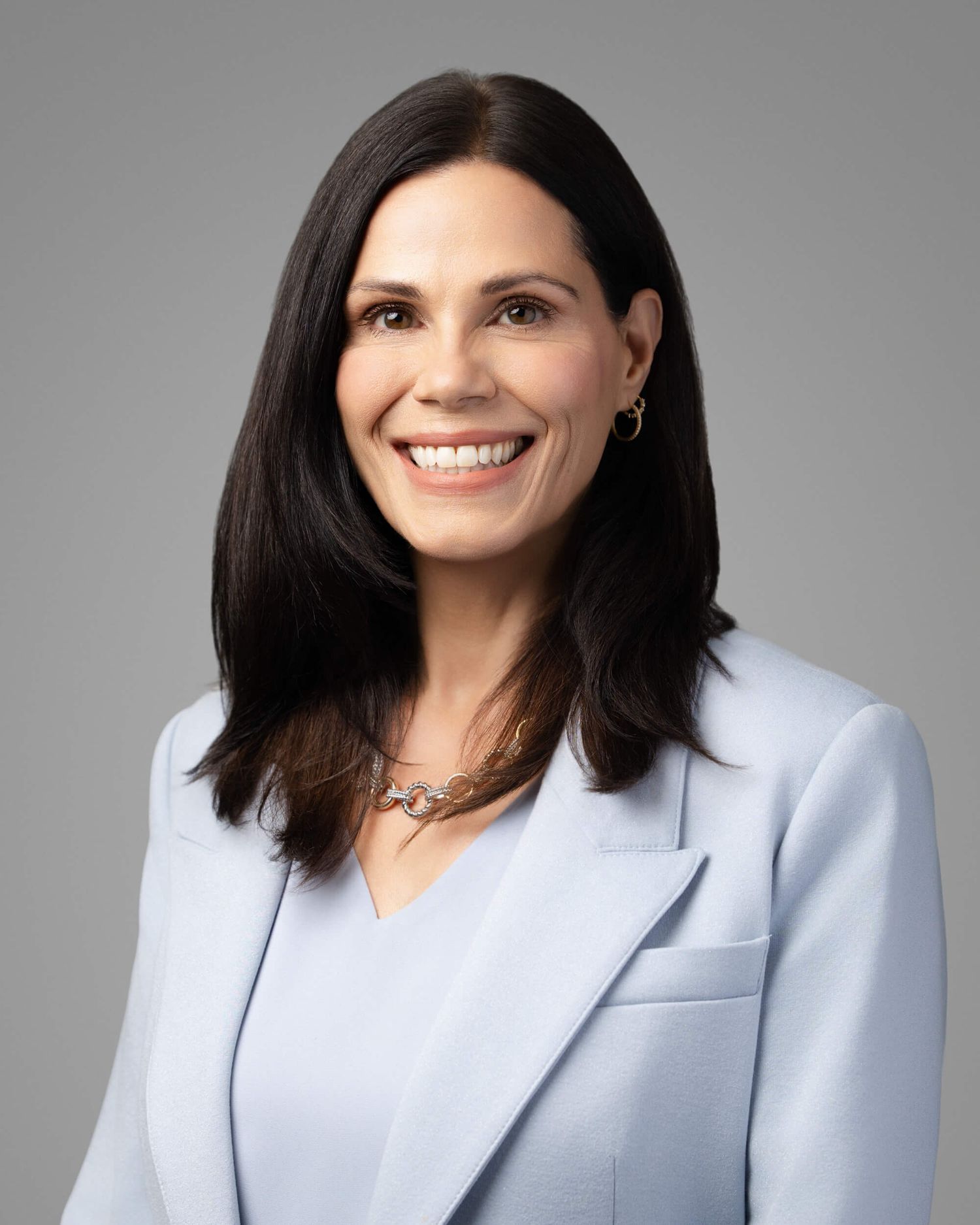 Professional headshot of a smiling woman with dark hair wearing a light blue blazer and gold necklace.