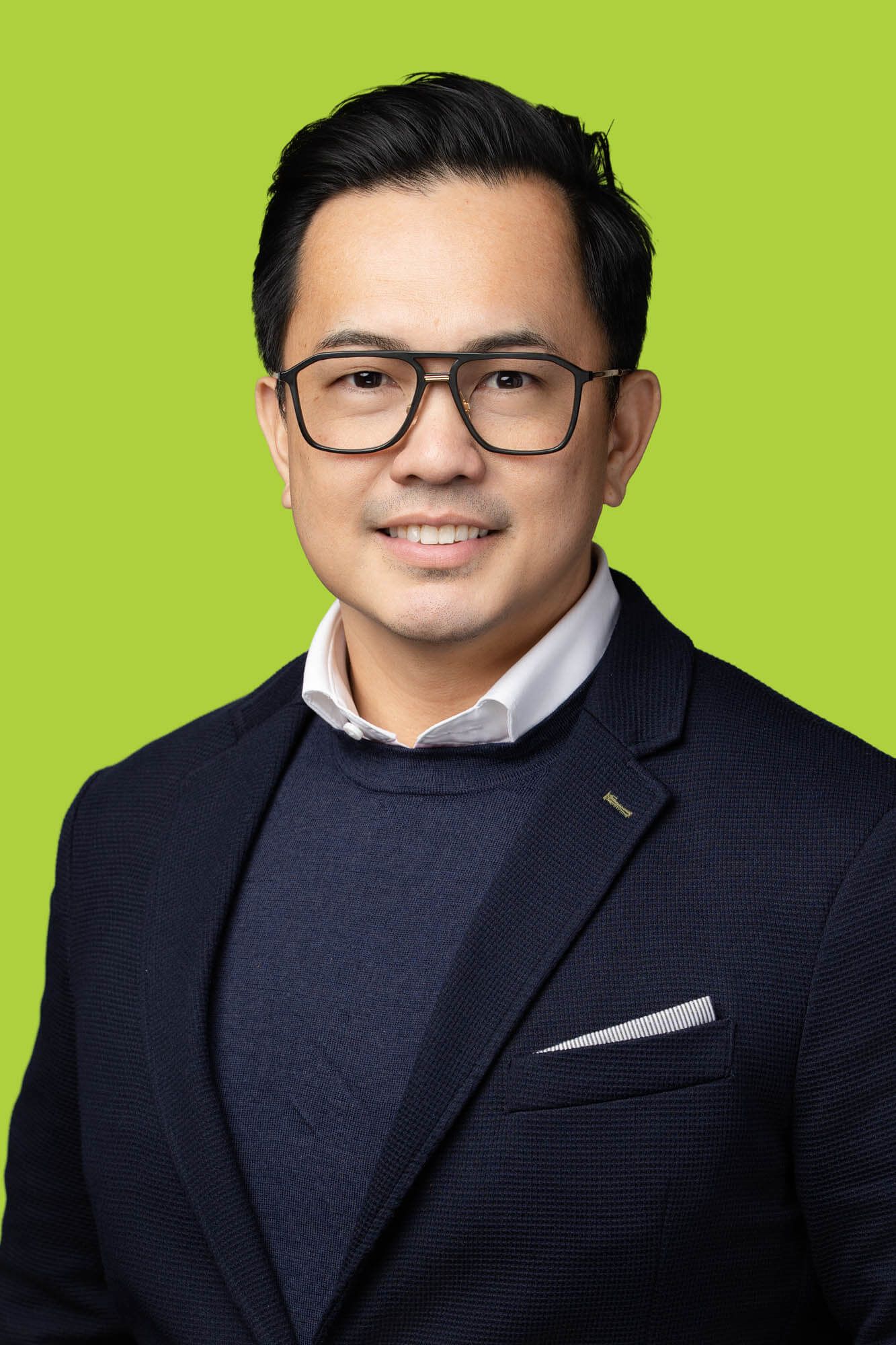 Professional headshot of a smiling Asian man wearing glasses and navy blazer against bright green background.