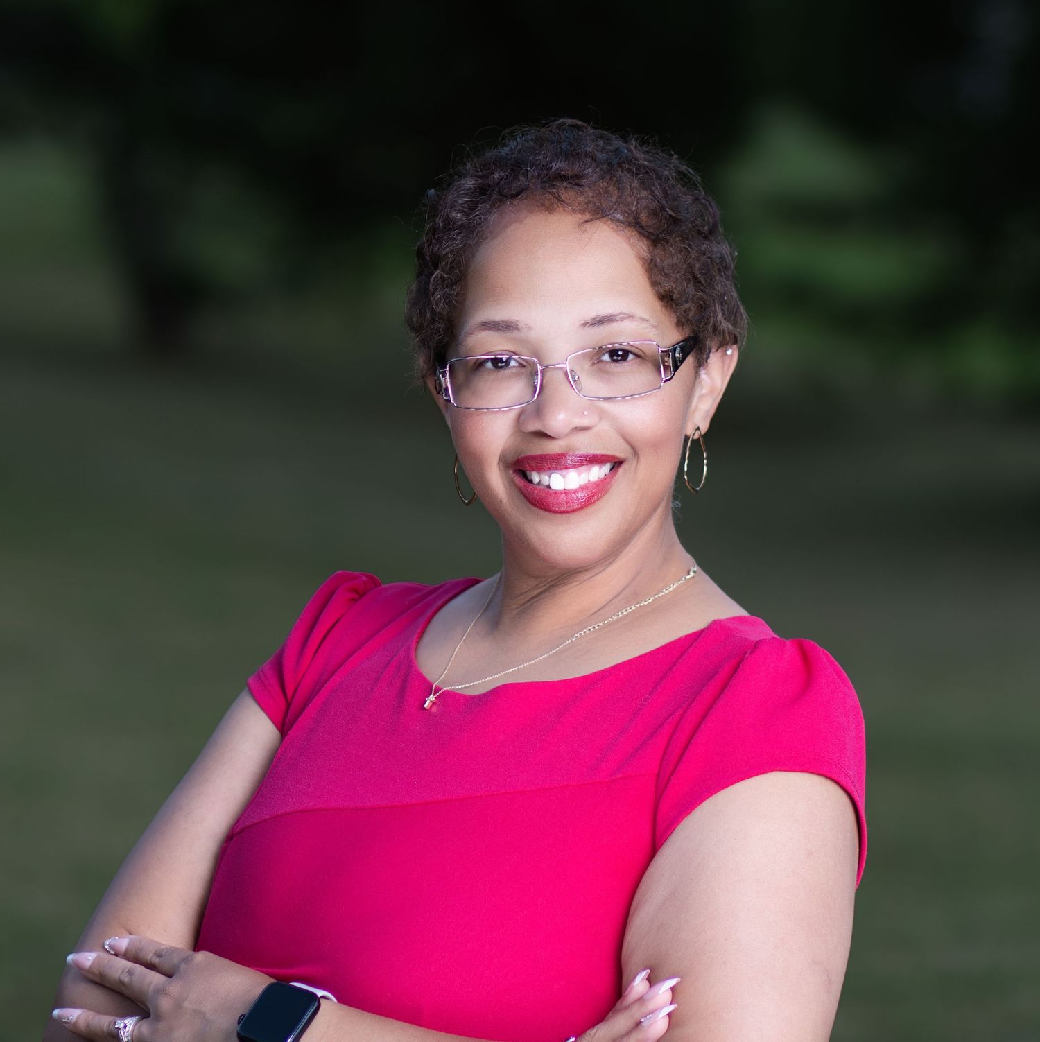 Professional woman in pink top with glasses, arms crossed, smiling outdoors in a corporate headshot photo.