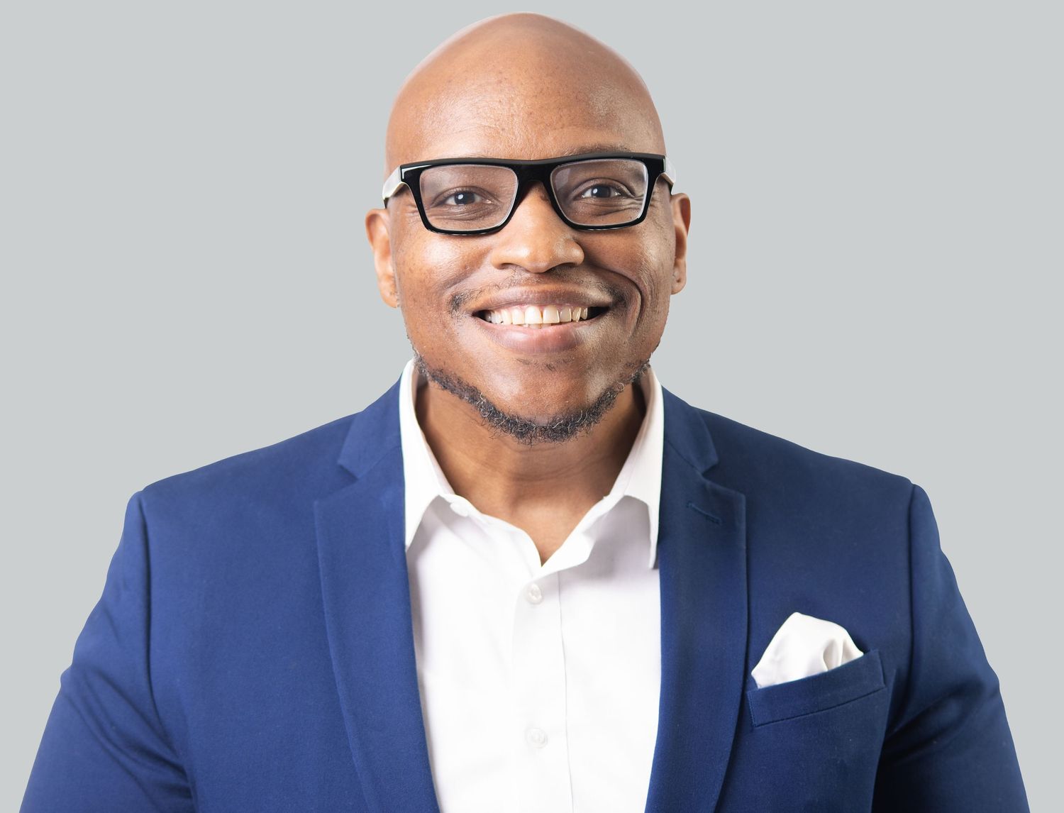 Smiling bald Black man wearing glasses and a blue blazer with white shirt, professional headshot on gray background.