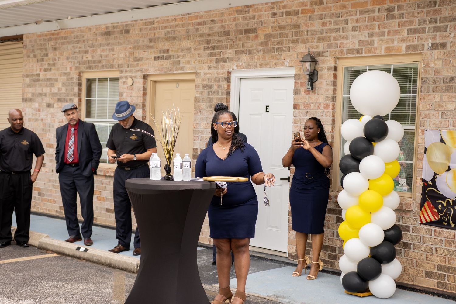 Ribbon cutting ceremony outside brick building with black, white, and yellow balloon decorations marking a grand opening.