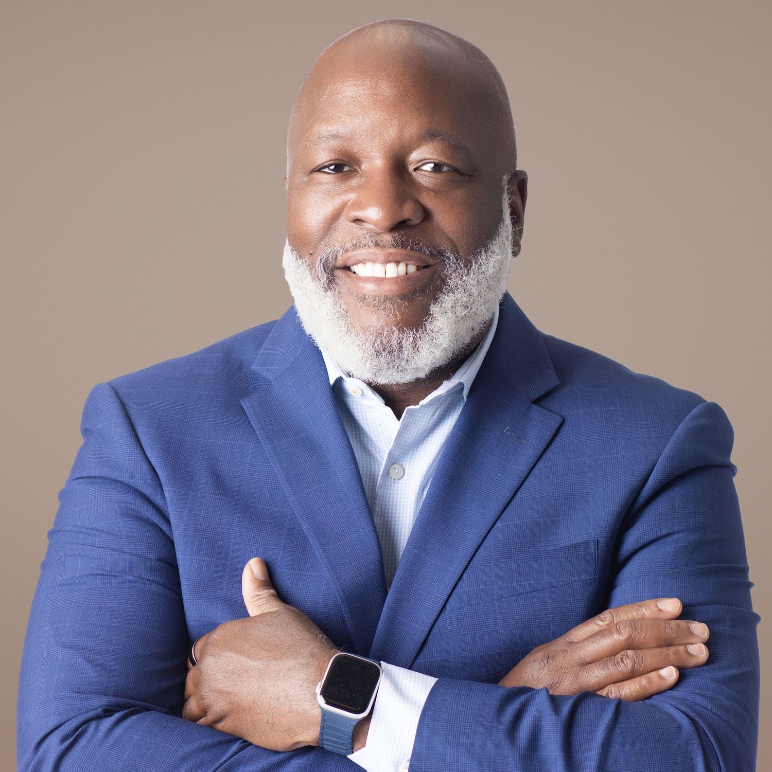 Confident smiling man in blue blazer with crossed arms, wearing a smartwatch, against a neutral beige background.
