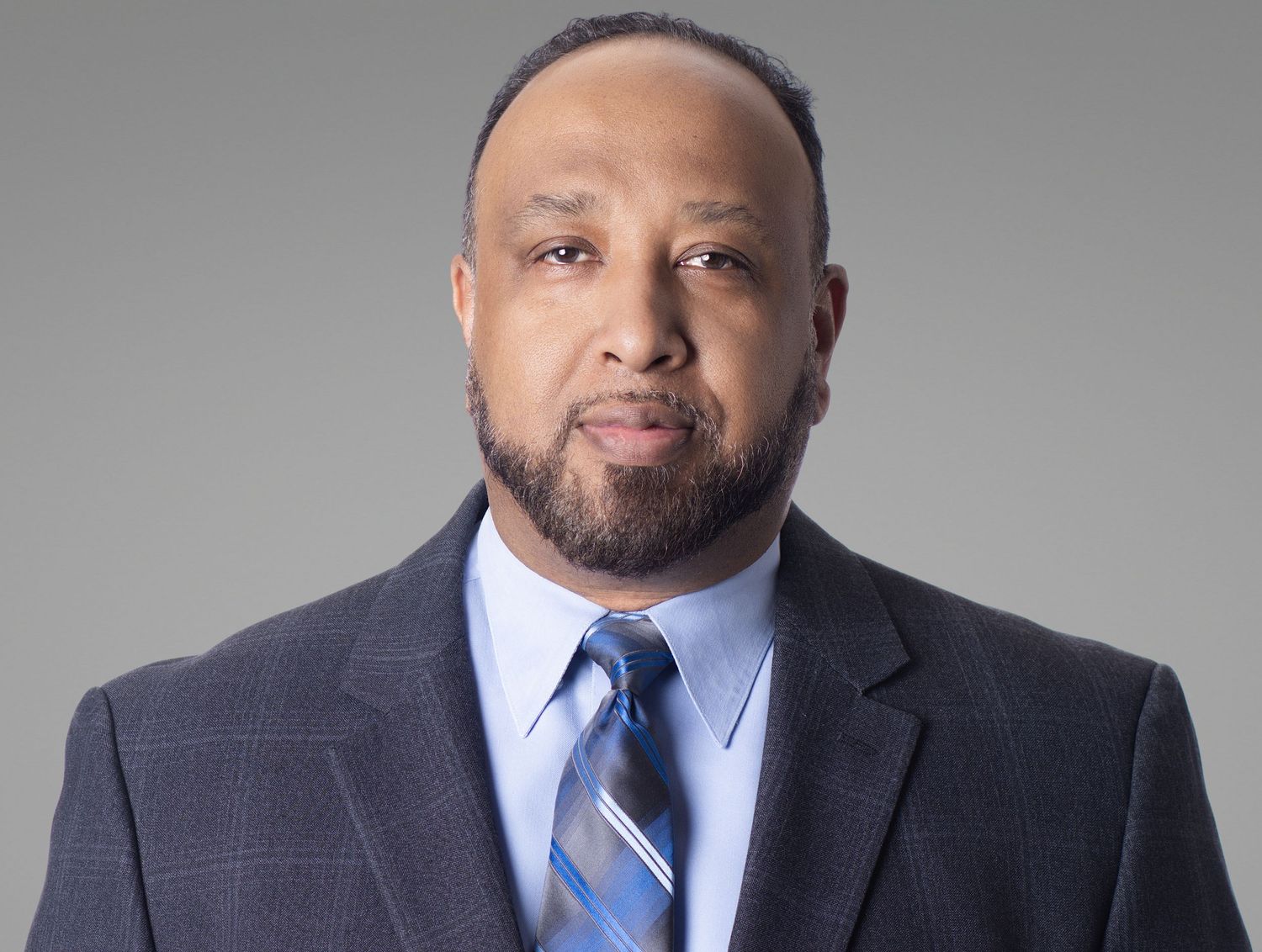 Professional man in a dark blazer and striped tie poses for a formal portrait against a gray background.