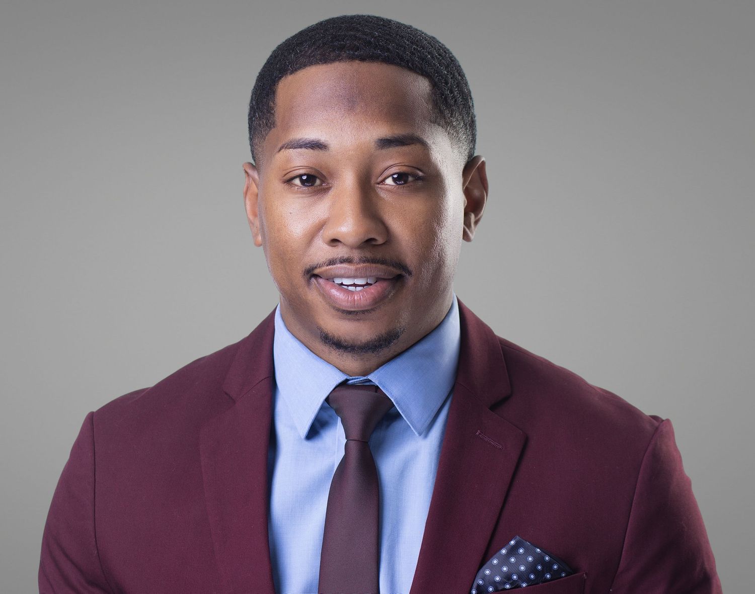 Professional man in maroon suit and blue shirt with polka dot pocket square poses for a formal headshot photo.