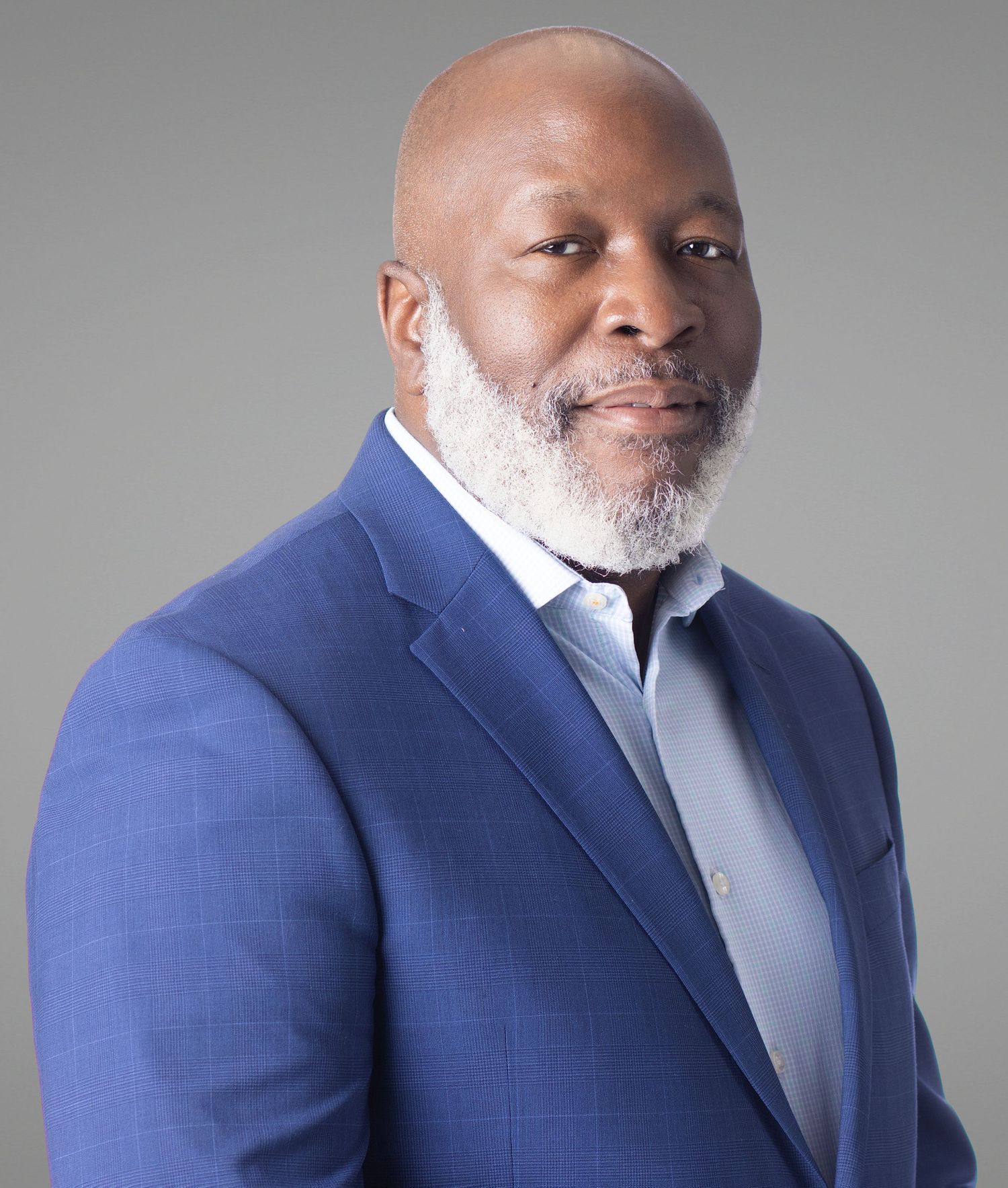 Professional headshot of a confident bald Black man with a white beard wearing a blue blazer against a gray background.