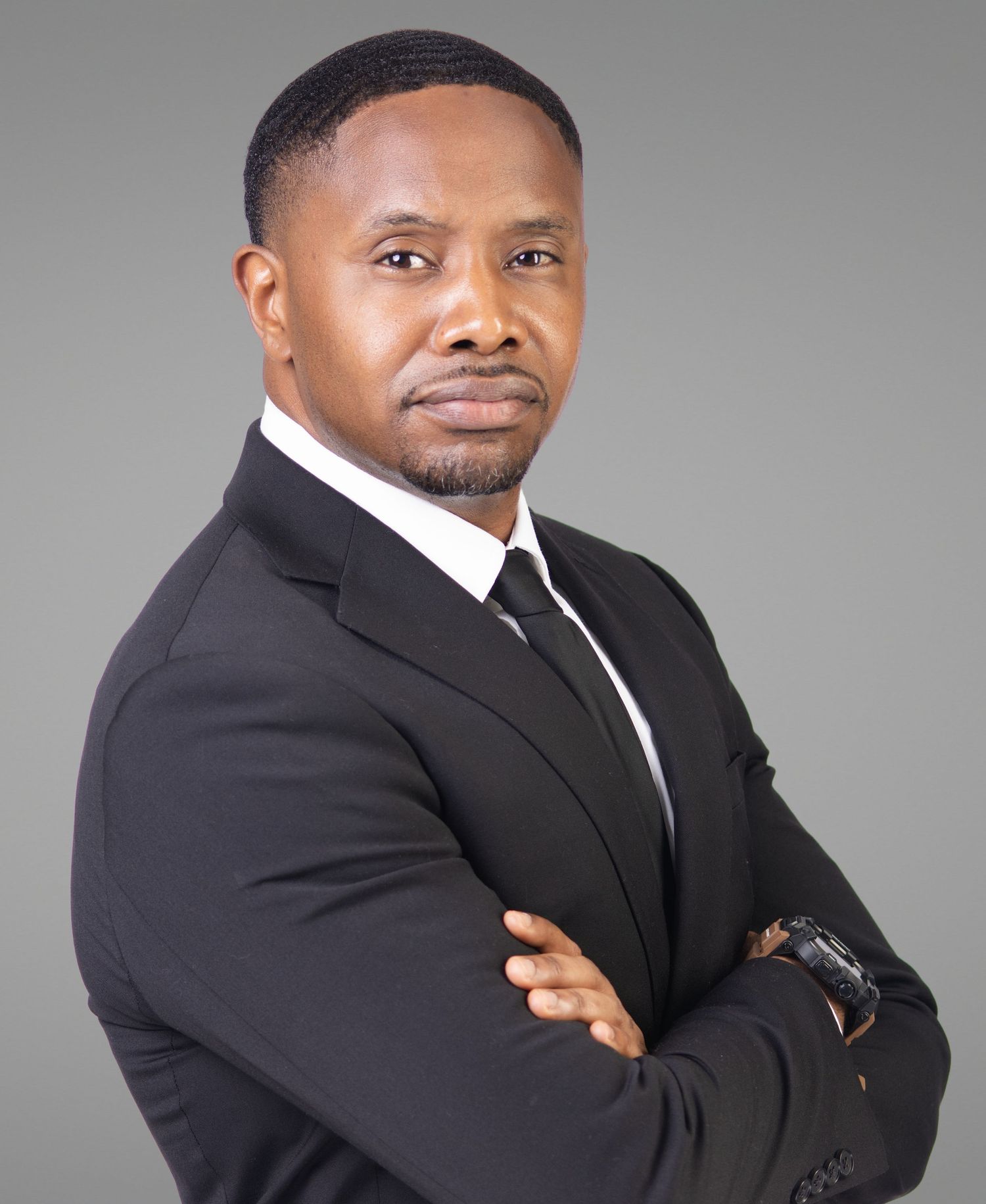 Professional man in black suit with arms crossed posing for a formal business portrait on gray background.
