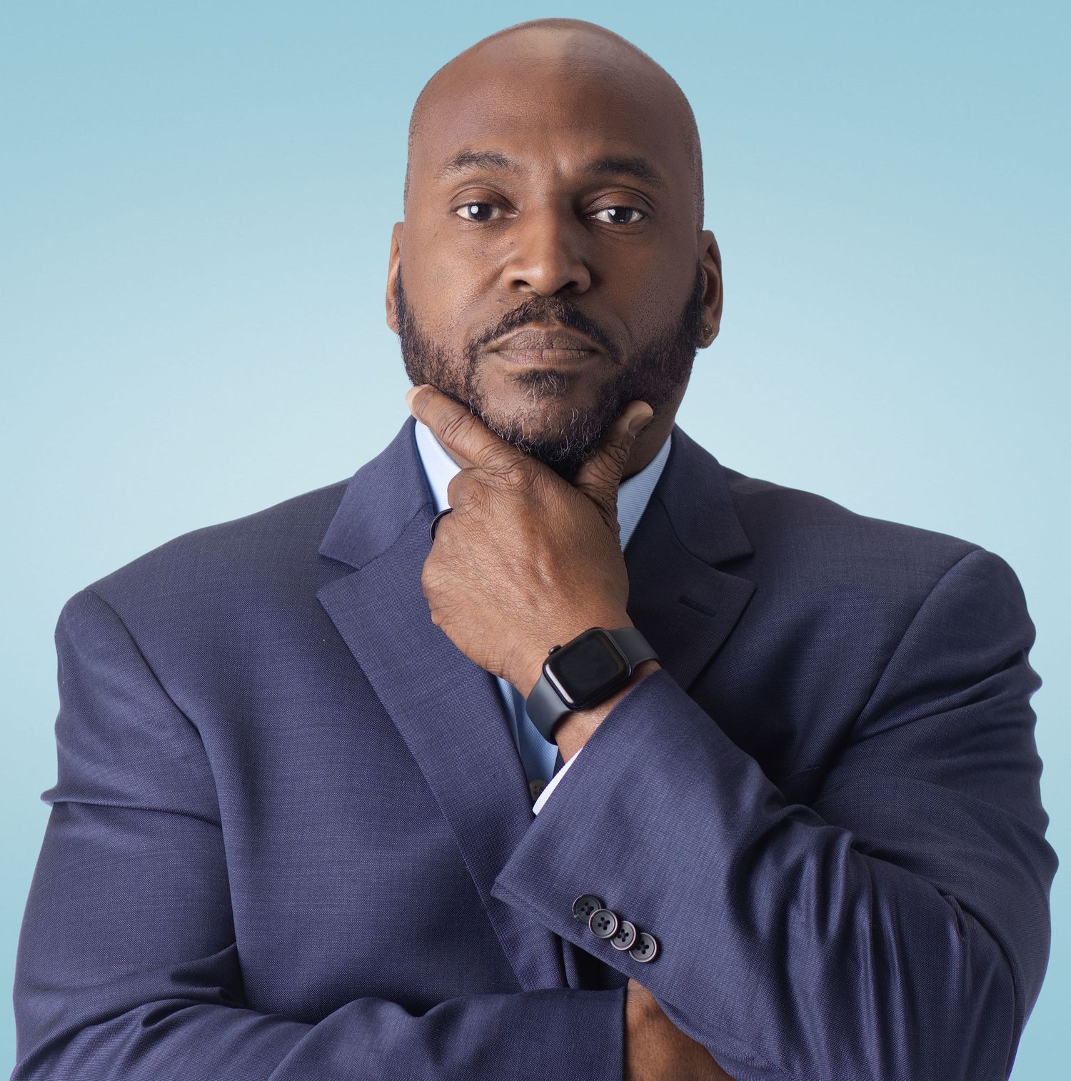 Professional Black man in navy suit, hand on chin, thoughtful pose against light blue background.
