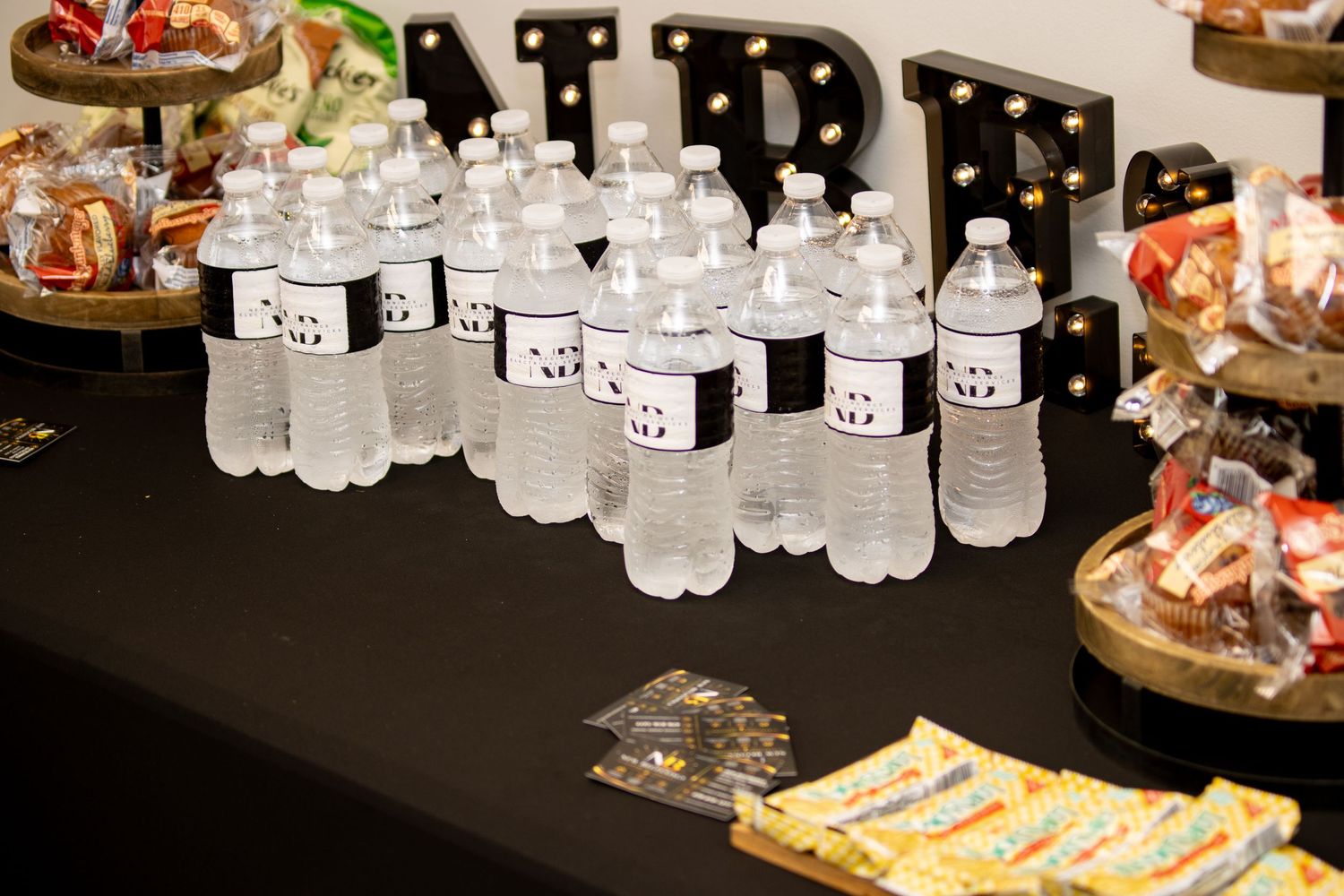 Water bottles with custom labels displayed on a black table at a party event with decorative marquee letters.