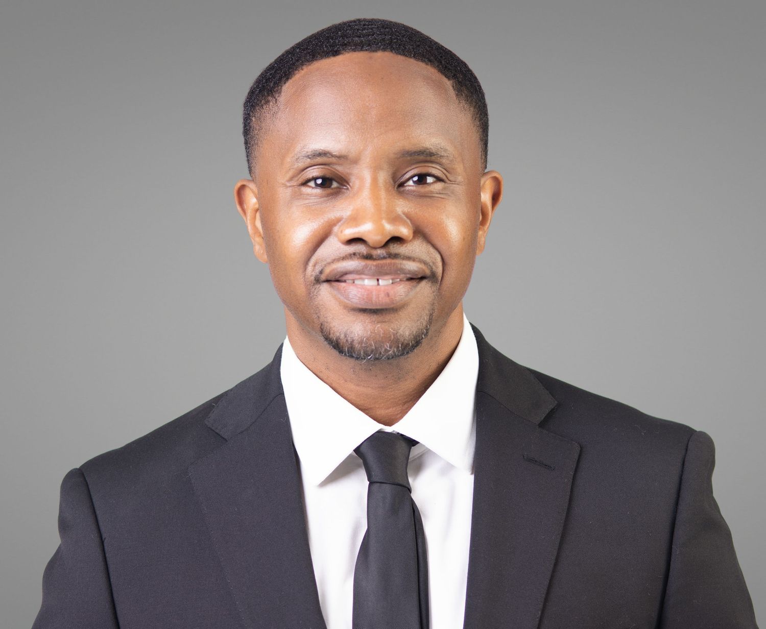 Professional headshot of a confident man in a black suit and tie against a gray background.