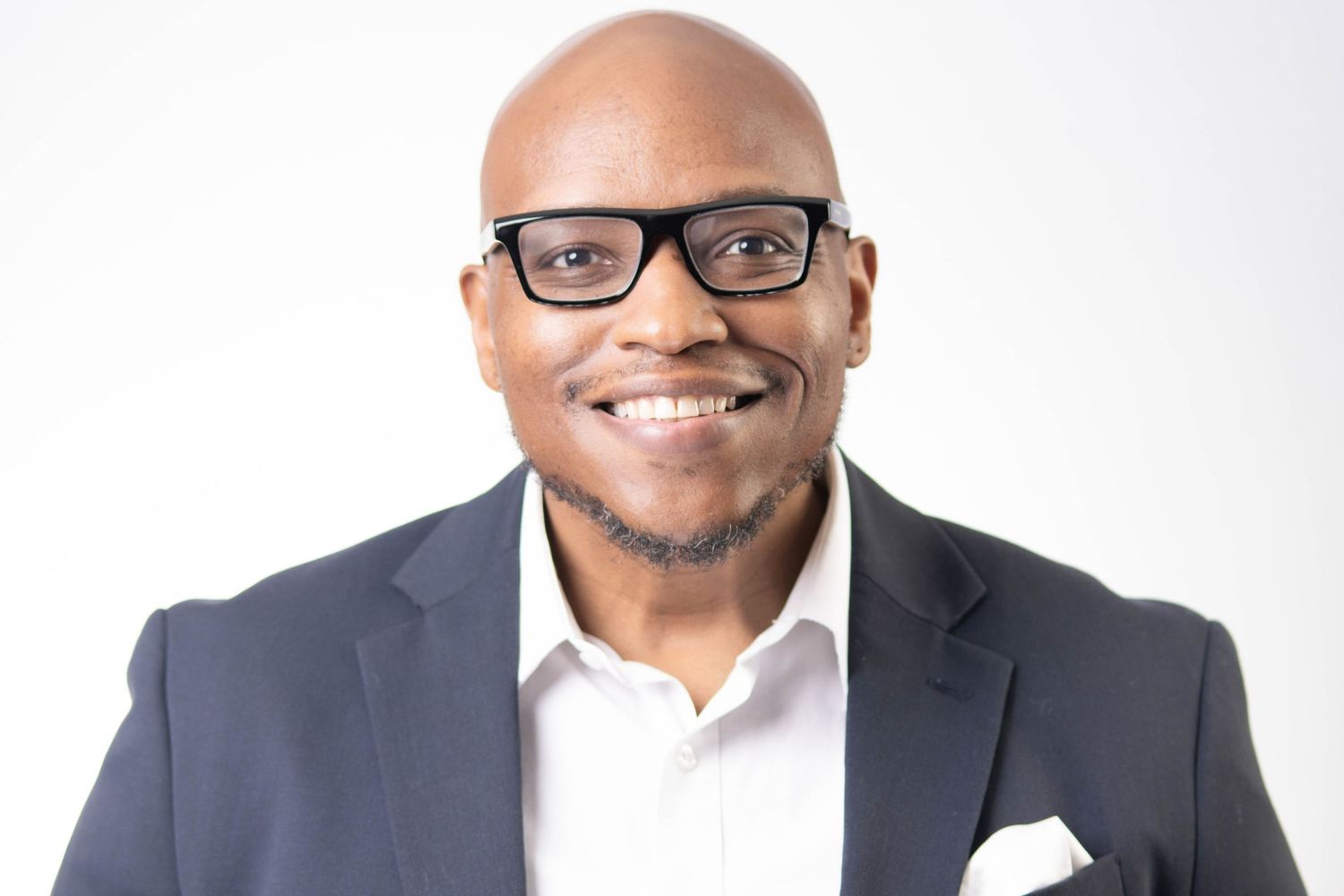 Smiling Black man in black-rimmed glasses and navy blazer poses for professional headshot on white background.