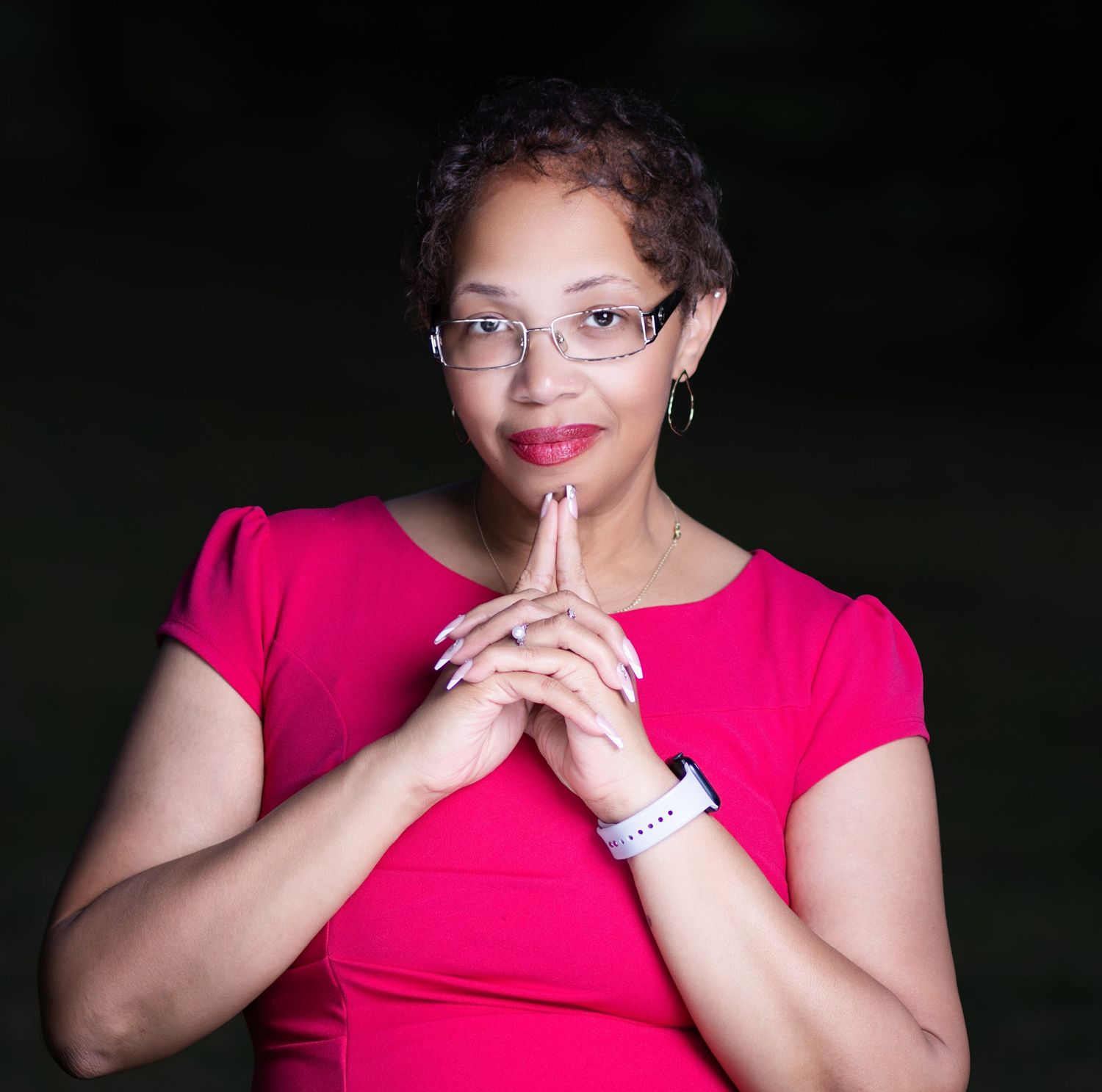 A confident woman in a bright pink dress with glasses poses with hands clasped, smiling against a dark background.