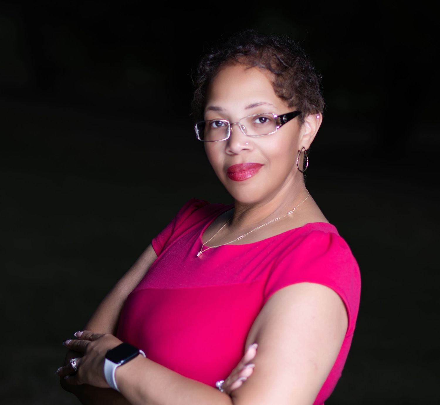 Professional woman in pink top and glasses poses confidently with arms crossed against dark background.