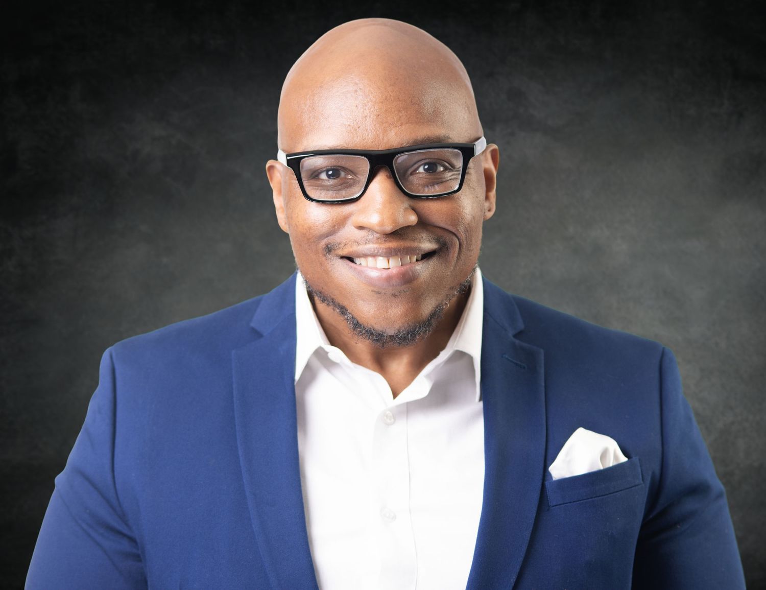 Professional man in blue blazer and glasses smiling confidently against dark background in formal portrait.