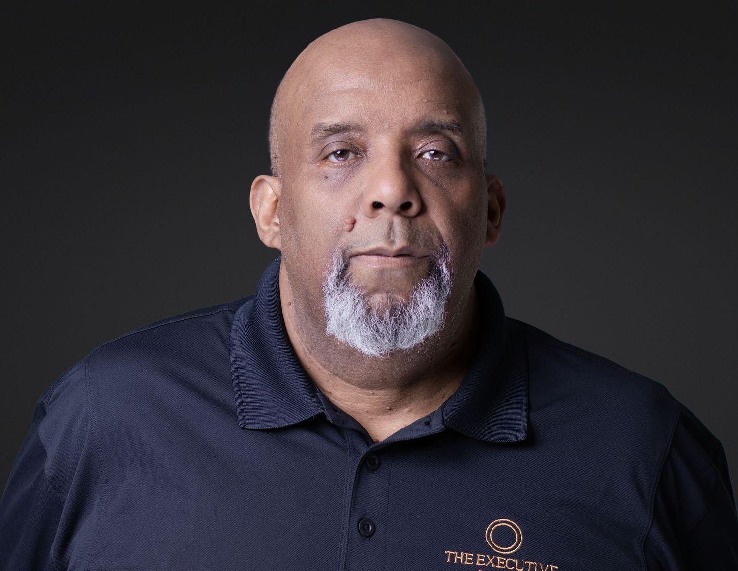 Professional headshot of a bald man with a gray beard wearing a navy blue polo shirt with a company logo.