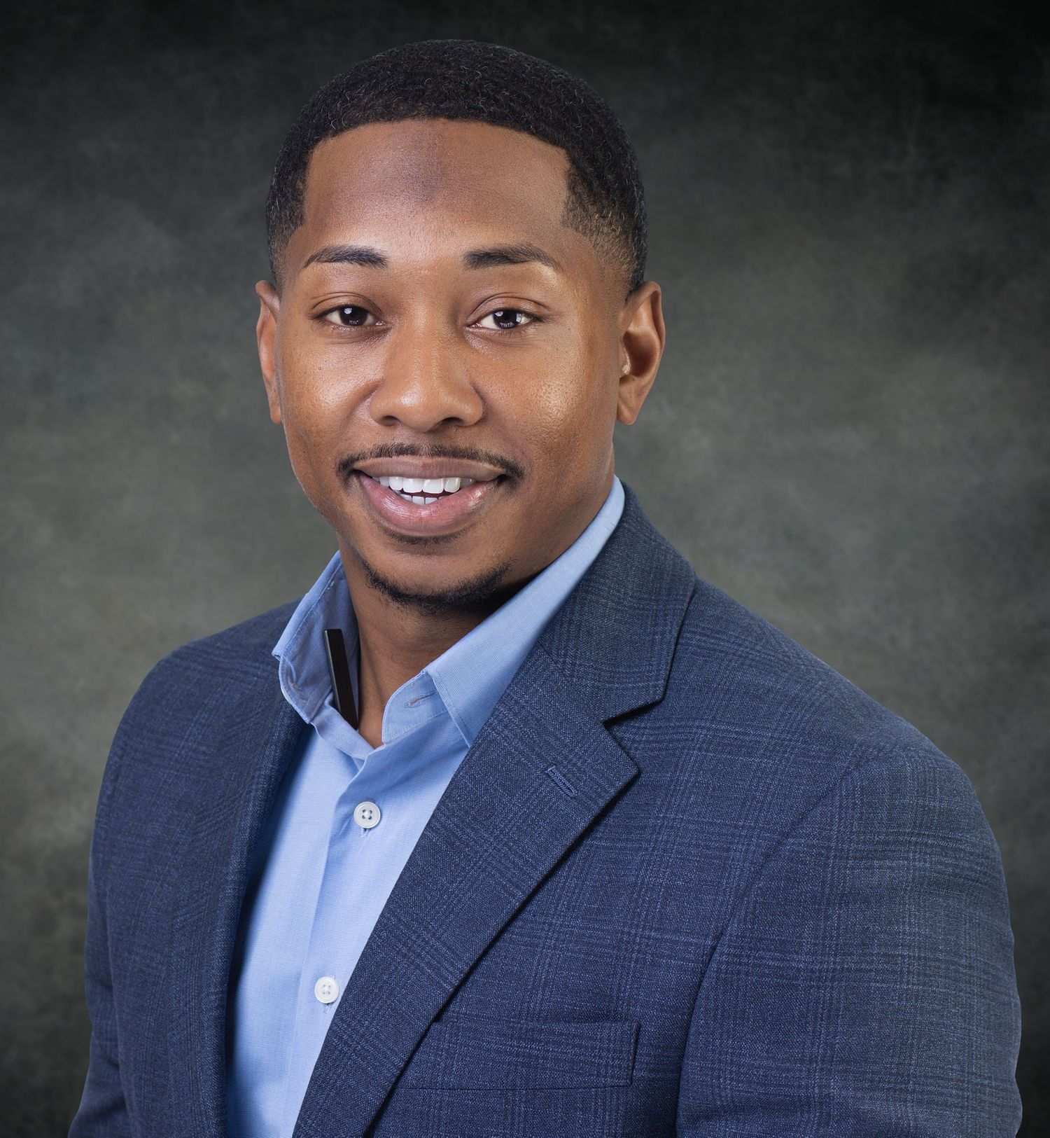 Professional headshot of a young man in a navy blazer and light blue shirt, smiling confidently against a gray background.