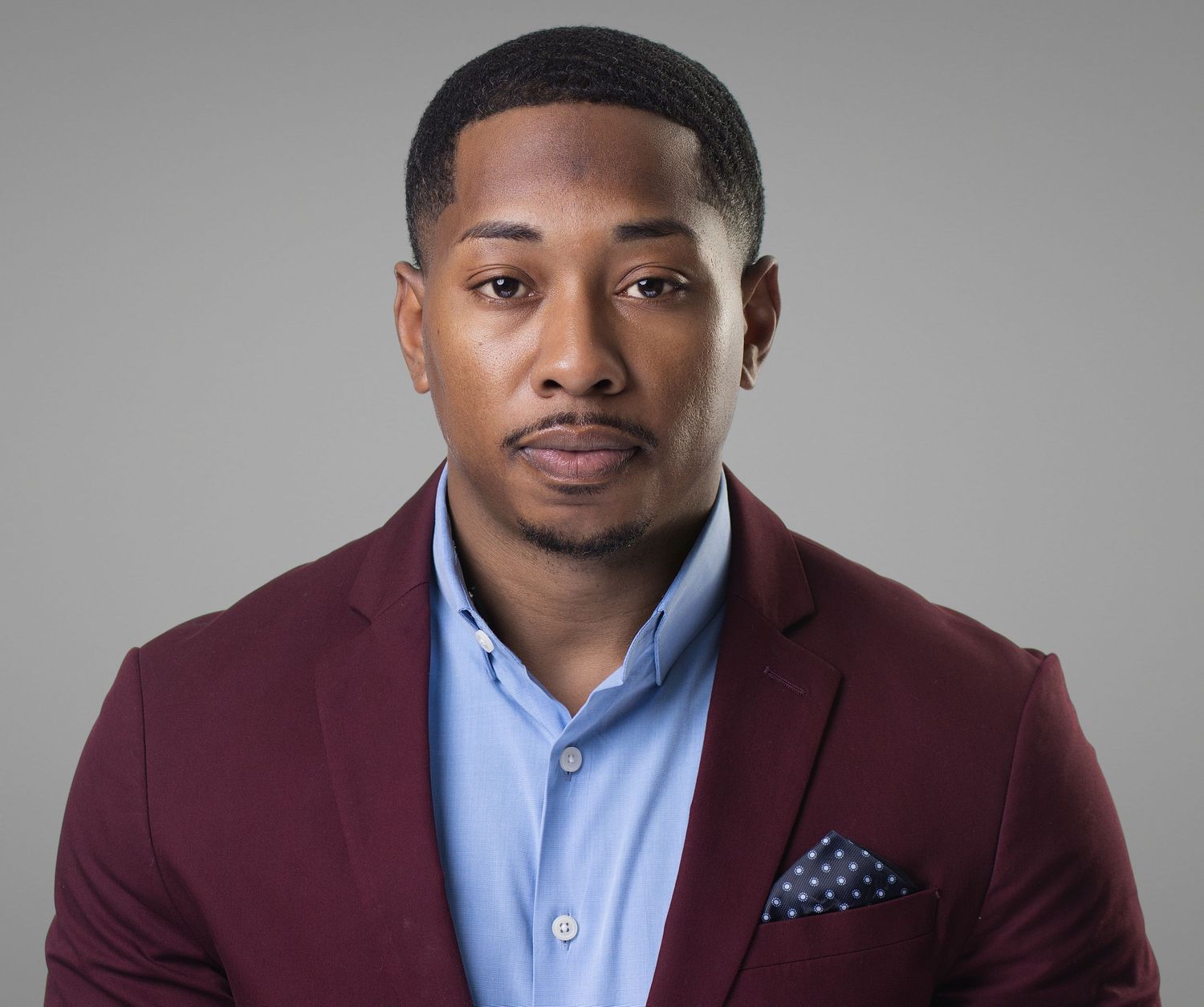 Professional headshot of a young Black man in a burgundy blazer and light blue shirt with pocket square.