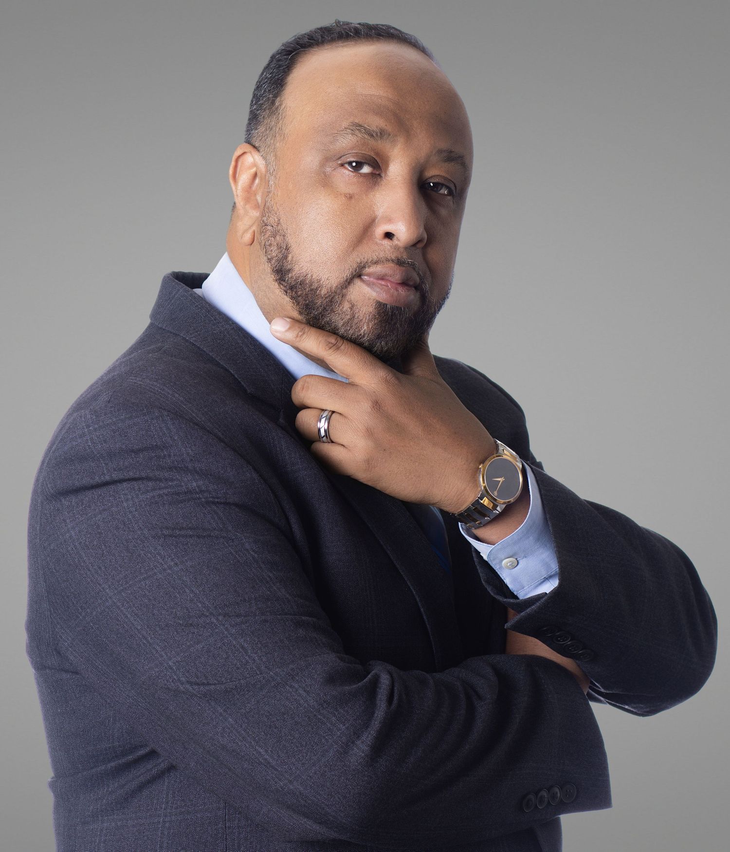 Professional man in dark suit with beard, posing thoughtfully with hand on chin against gray background.