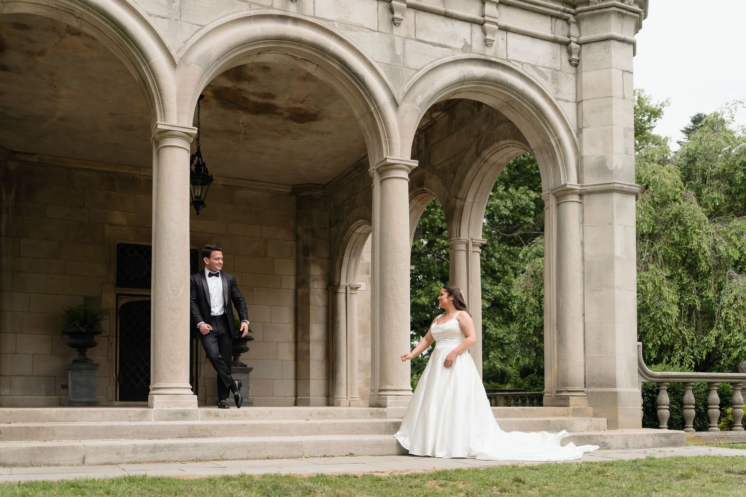 Bride and groom pose under stone arches of elegant historic venue surrounded by lush greenery on their wedding day.
