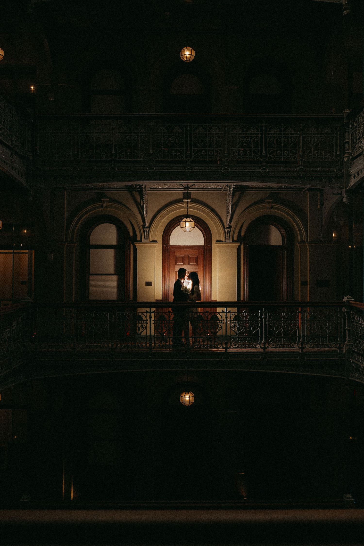 Silhouetted couple embracing on ornate balcony of elegant historic building illuminated by warm golden light at night.