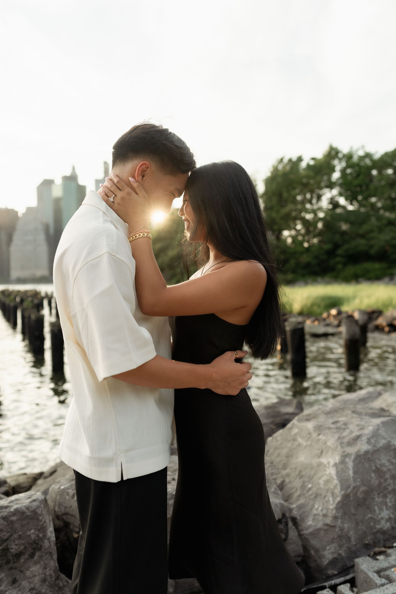 Couple embracing near waterfront with city skyline and golden sunlight glowing between them, wearing black and white outfits.
