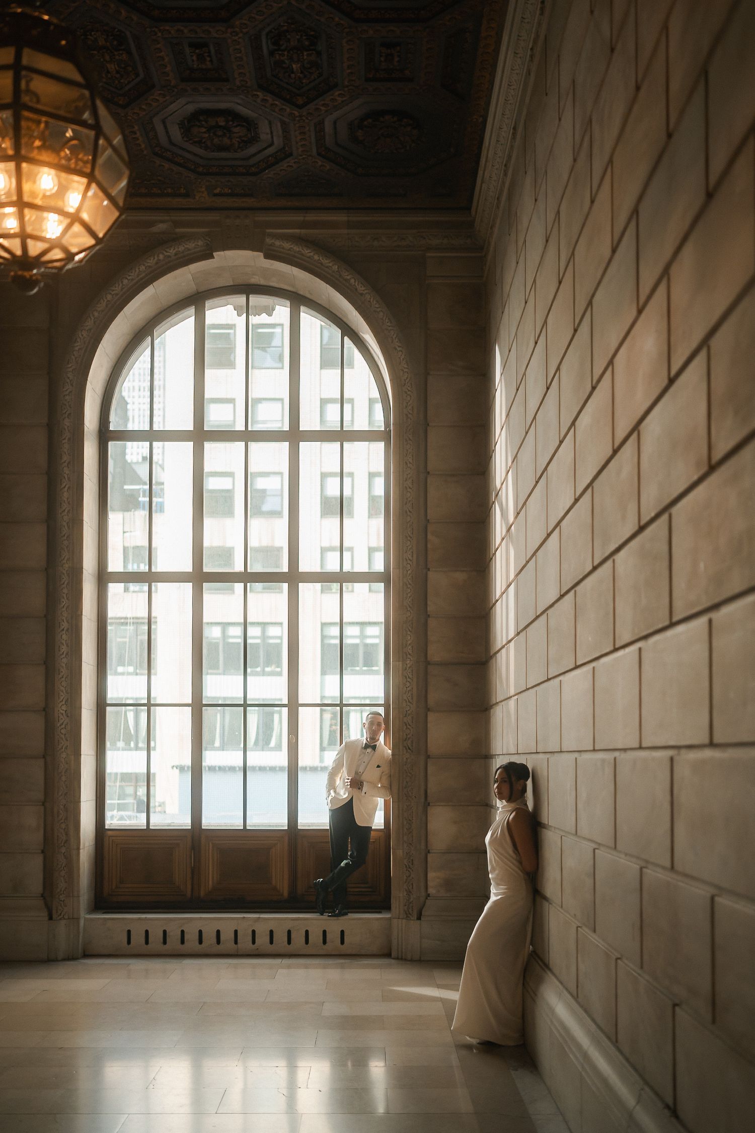 Couple poses in elegant stone hallway with arched window, chandelier, and city views in romantic engagement photo.
