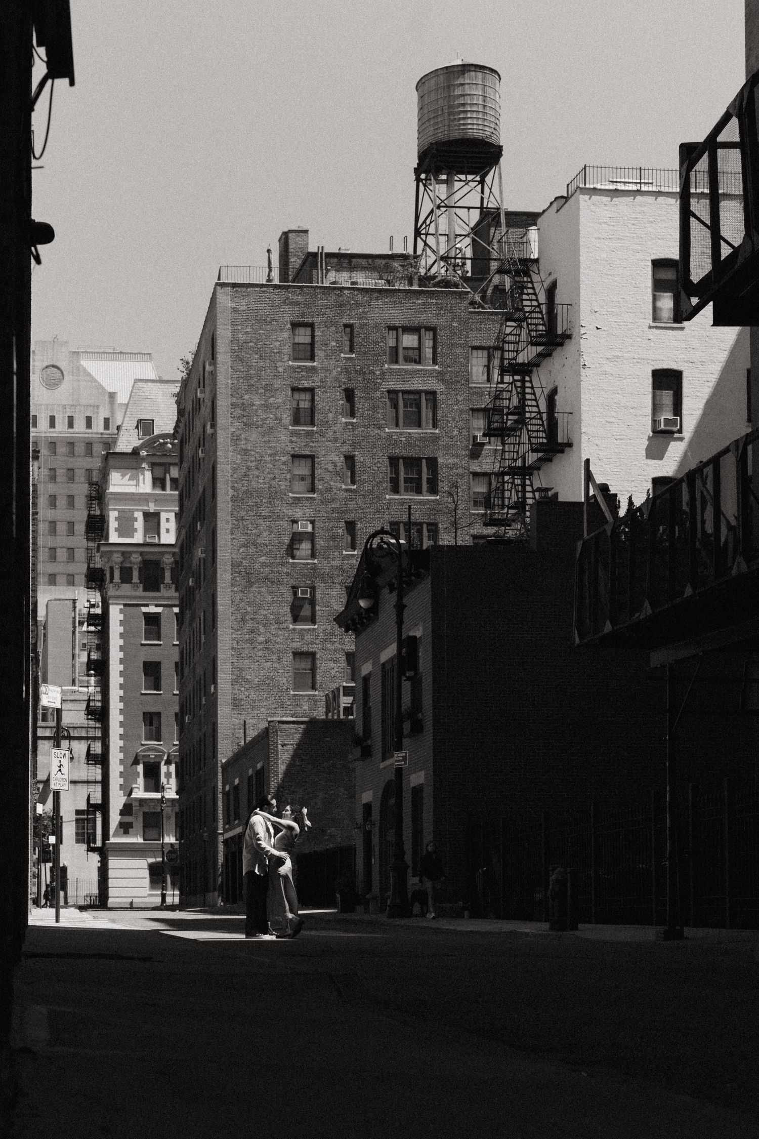 Black and white photo of a couple embracing on a shadowy NYC street with brick buildings and a rooftop water tower.
