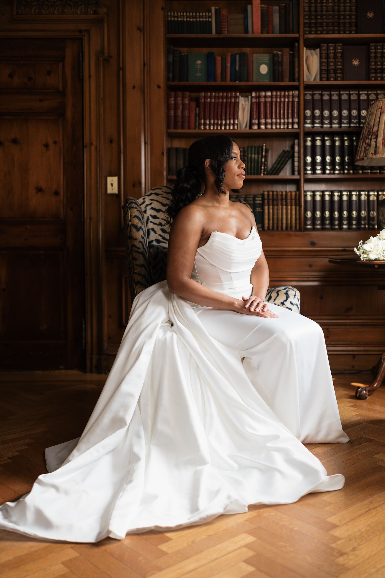 Elegant bride in a strapless white satin gown seated in a library chair, gazing thoughtfully to the side.