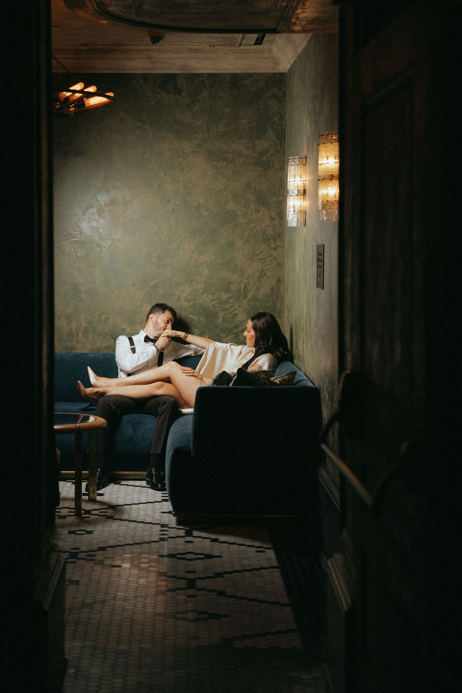 Couple relaxing on blue velvet sofas in a dimly lit, elegantly decorated lounge with green walls and warm lighting.