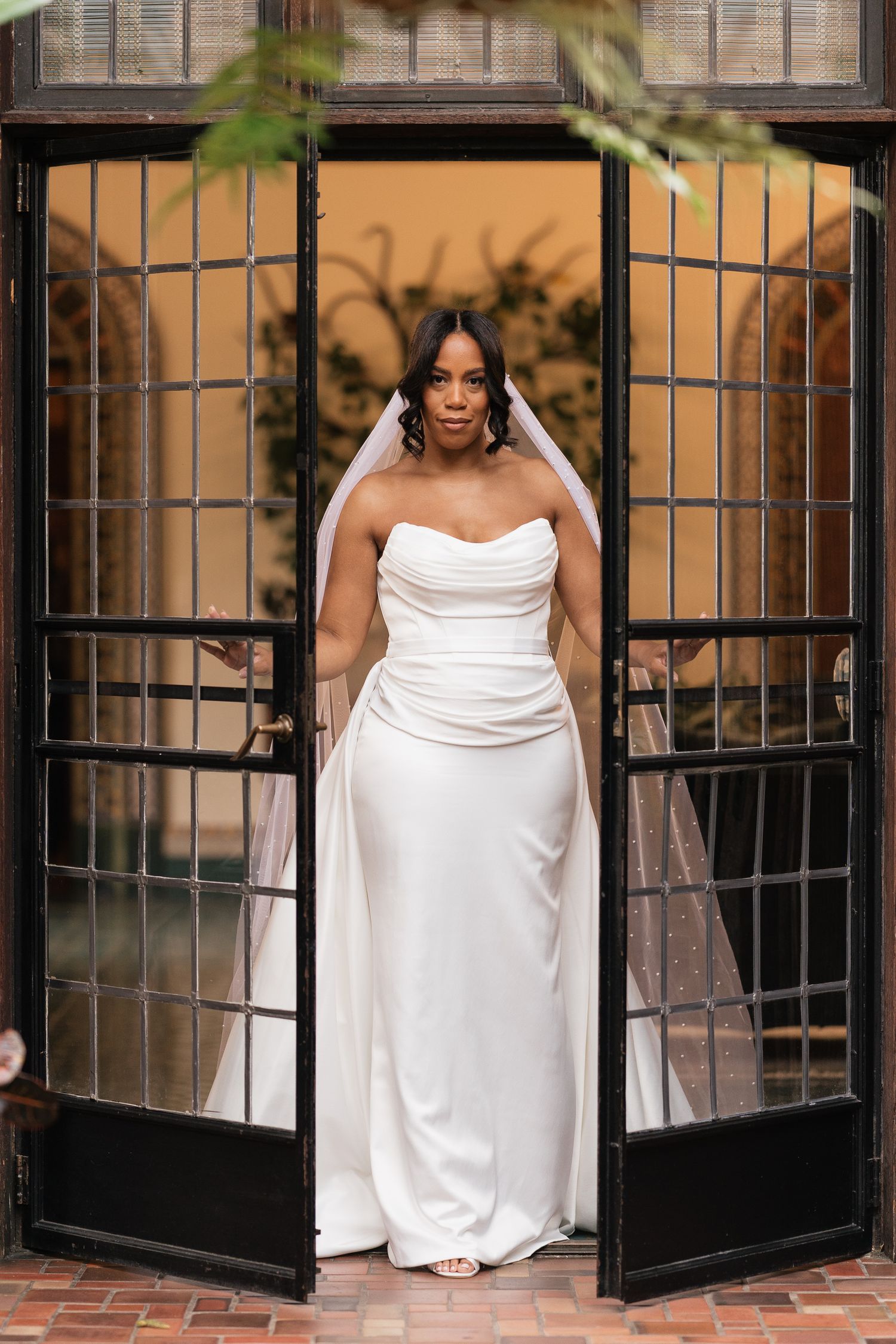 Elegant bride in strapless white gown and veil stands in black French doors at a wedding venue.