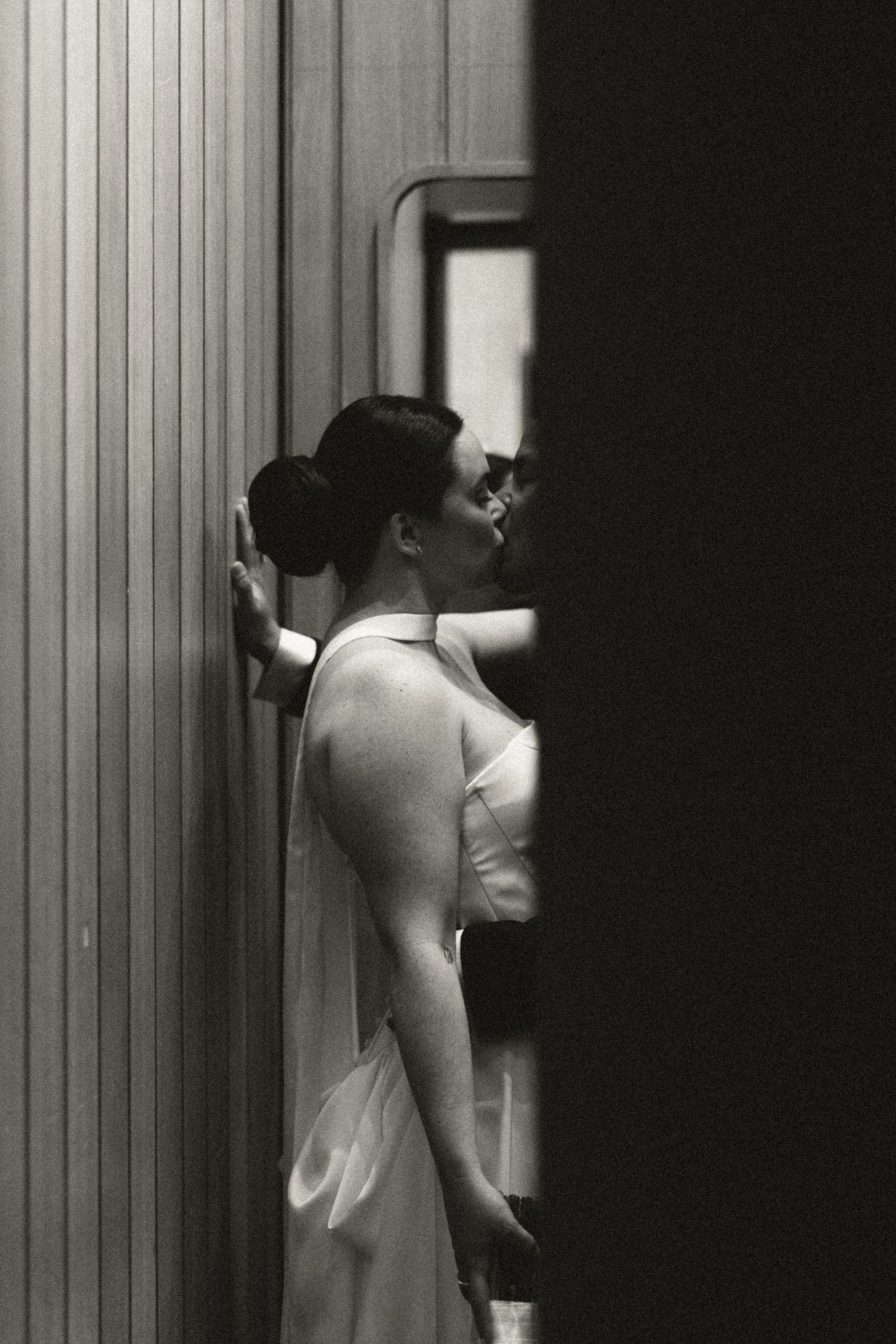 Black and white photo of a woman in a white dress leaning against a wall, viewed from the side.