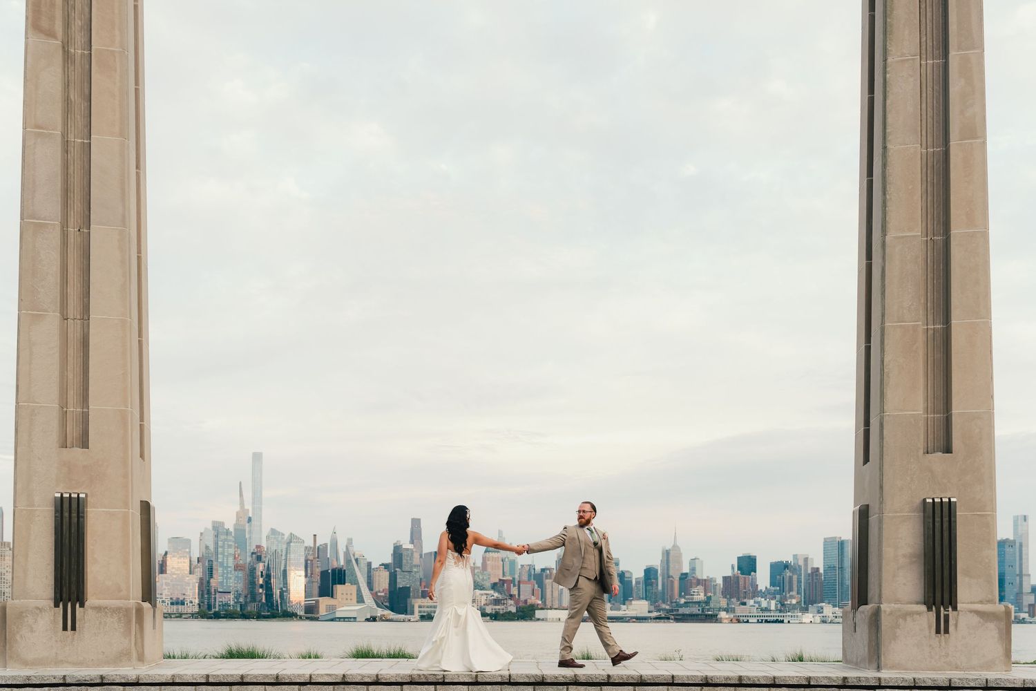 Bride and groom hold hands between grand columns with NYC skyline and waterfront backdrop on wedding day.