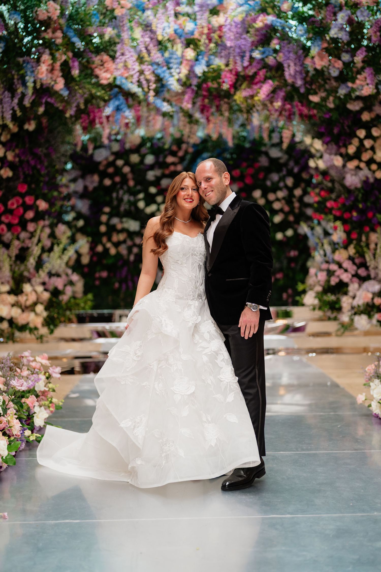 Bride in white ball gown and groom in black tuxedo pose under stunning floral arch with colorful blooms.