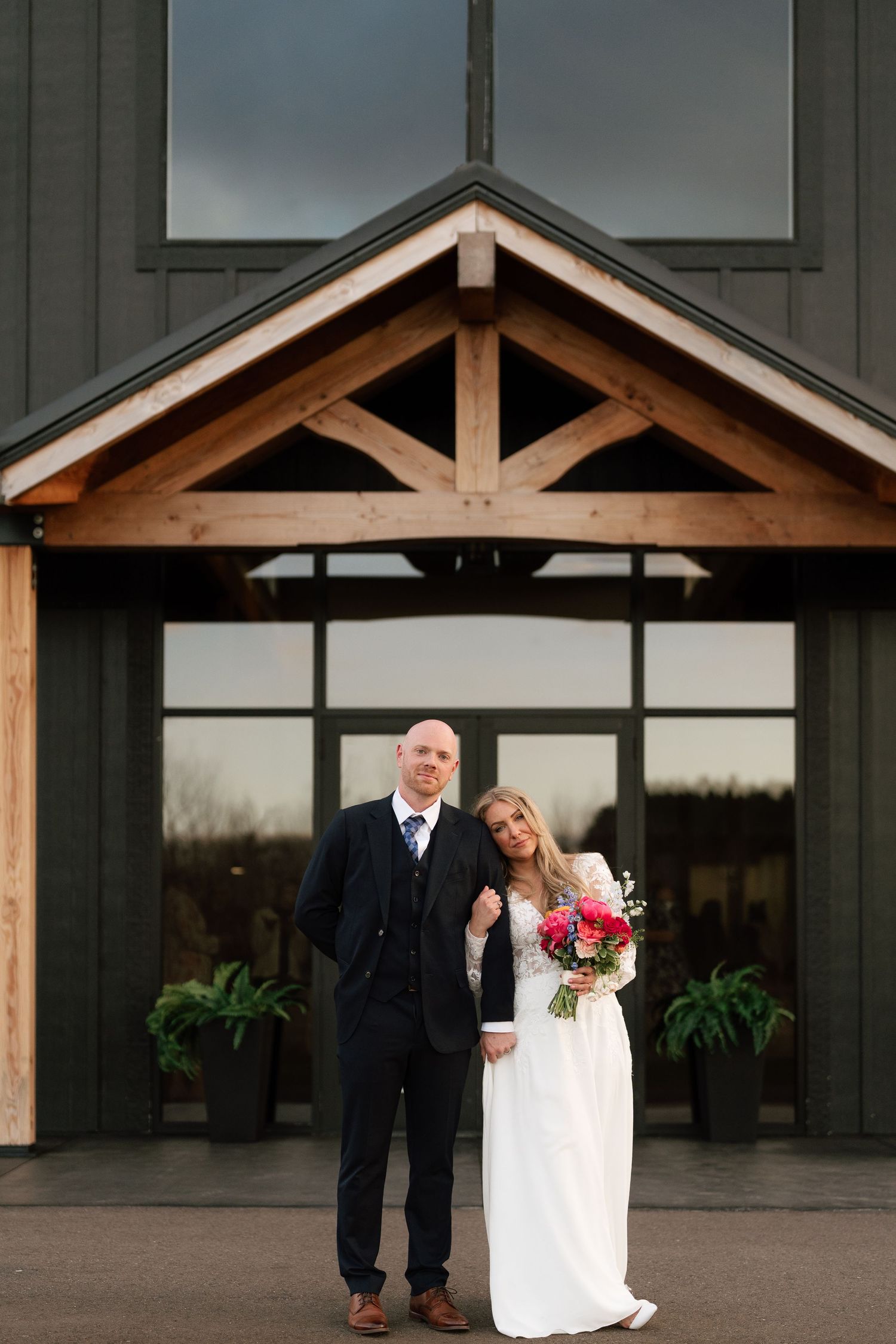 Bride and groom posing in front of a rustic barn venue with wooden beams, bride holding a vibrant pink bouquet.