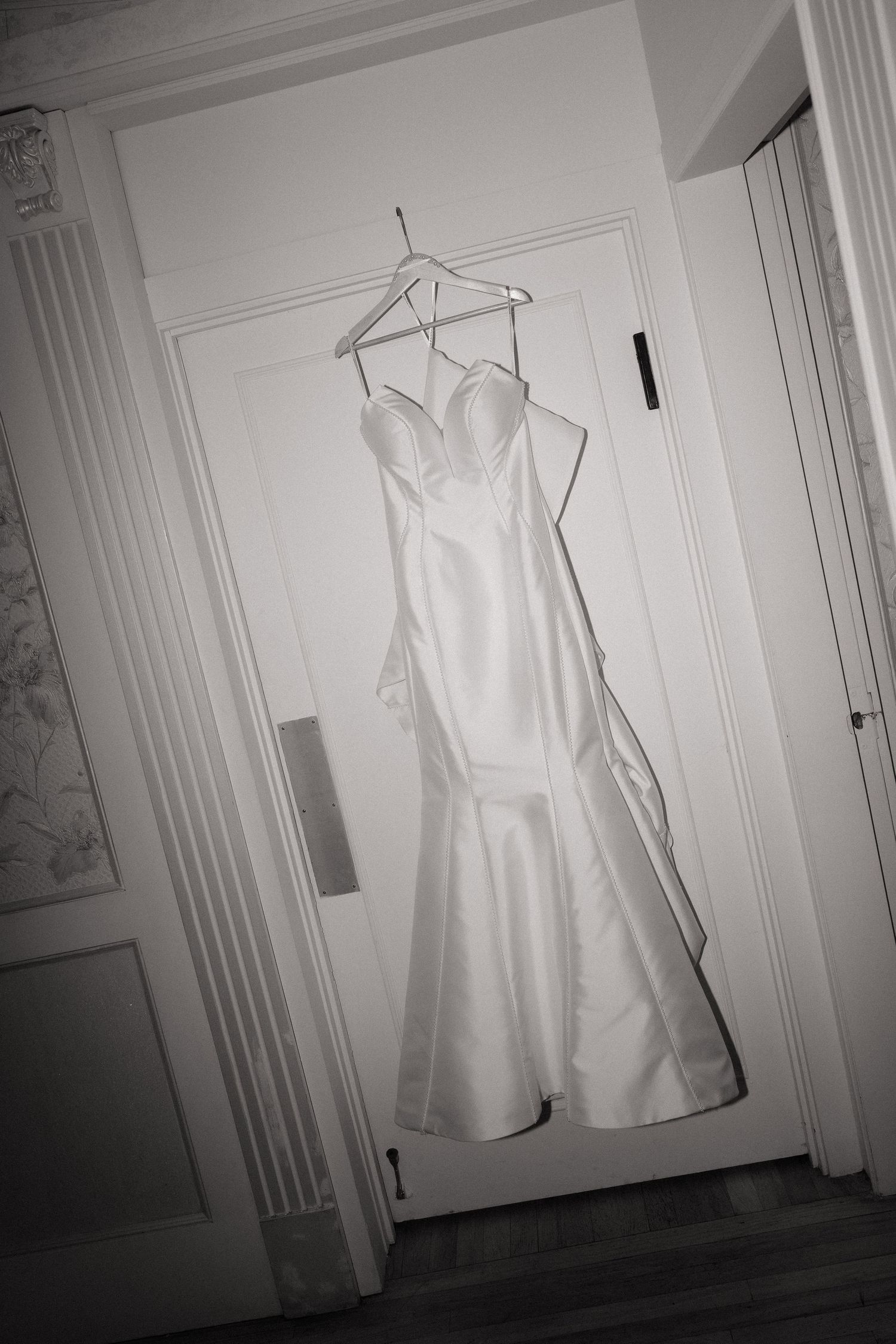 A white wedding dress with sweetheart neckline and mermaid silhouette hangs on a hanger against a white door.