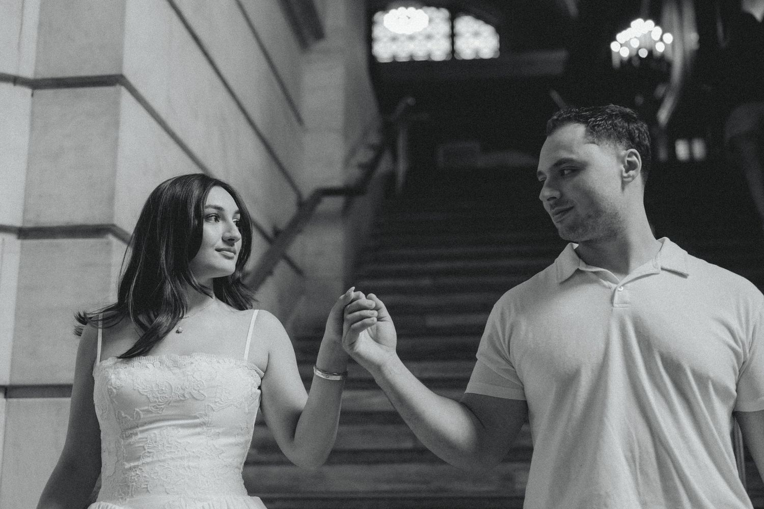 Black and white photo of a couple holding hands on grand staircase steps, woman in white dress, man in polo shirt.