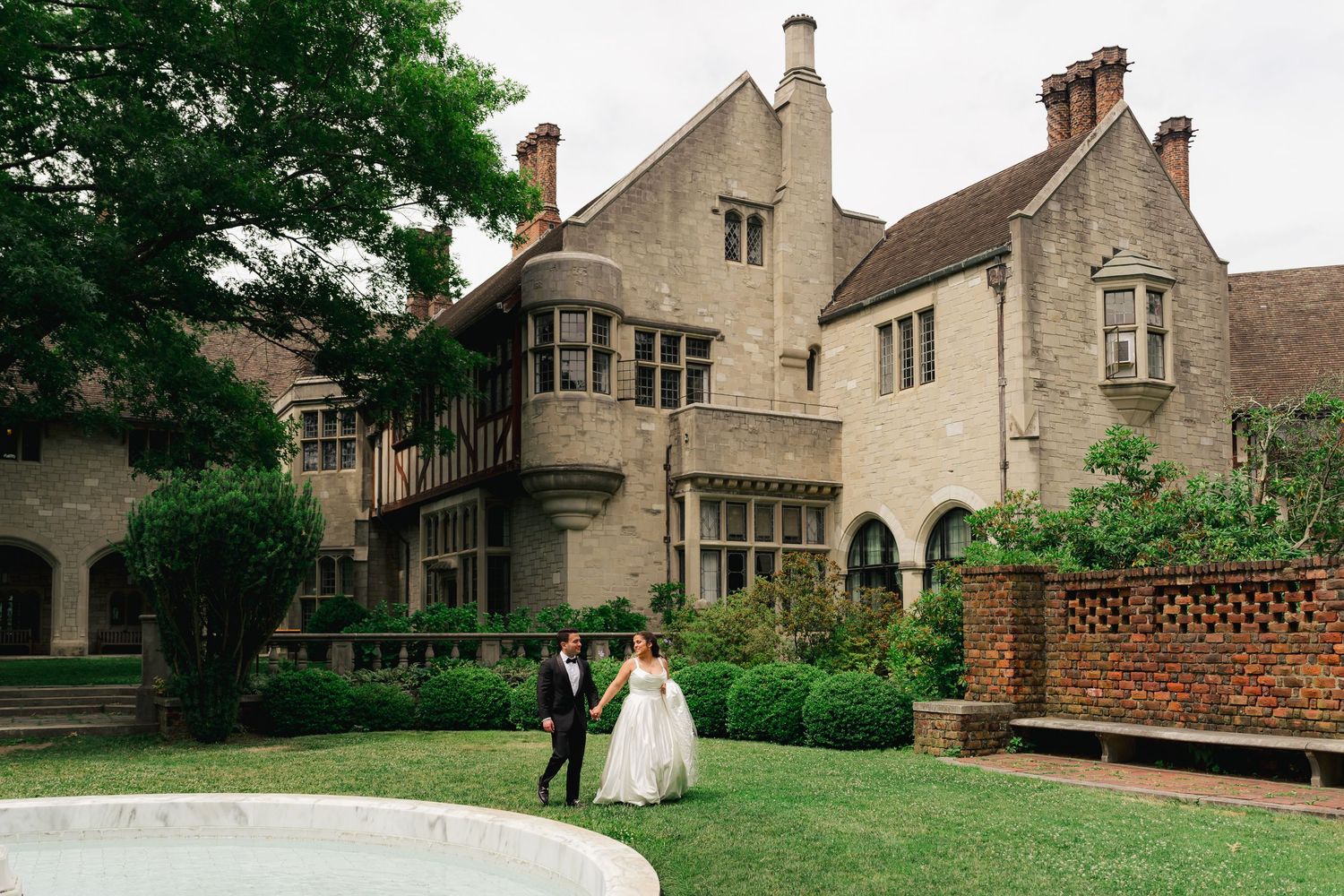 Bride and groom walking hand in hand on lush green lawn outside elegant stone manor wedding venue.