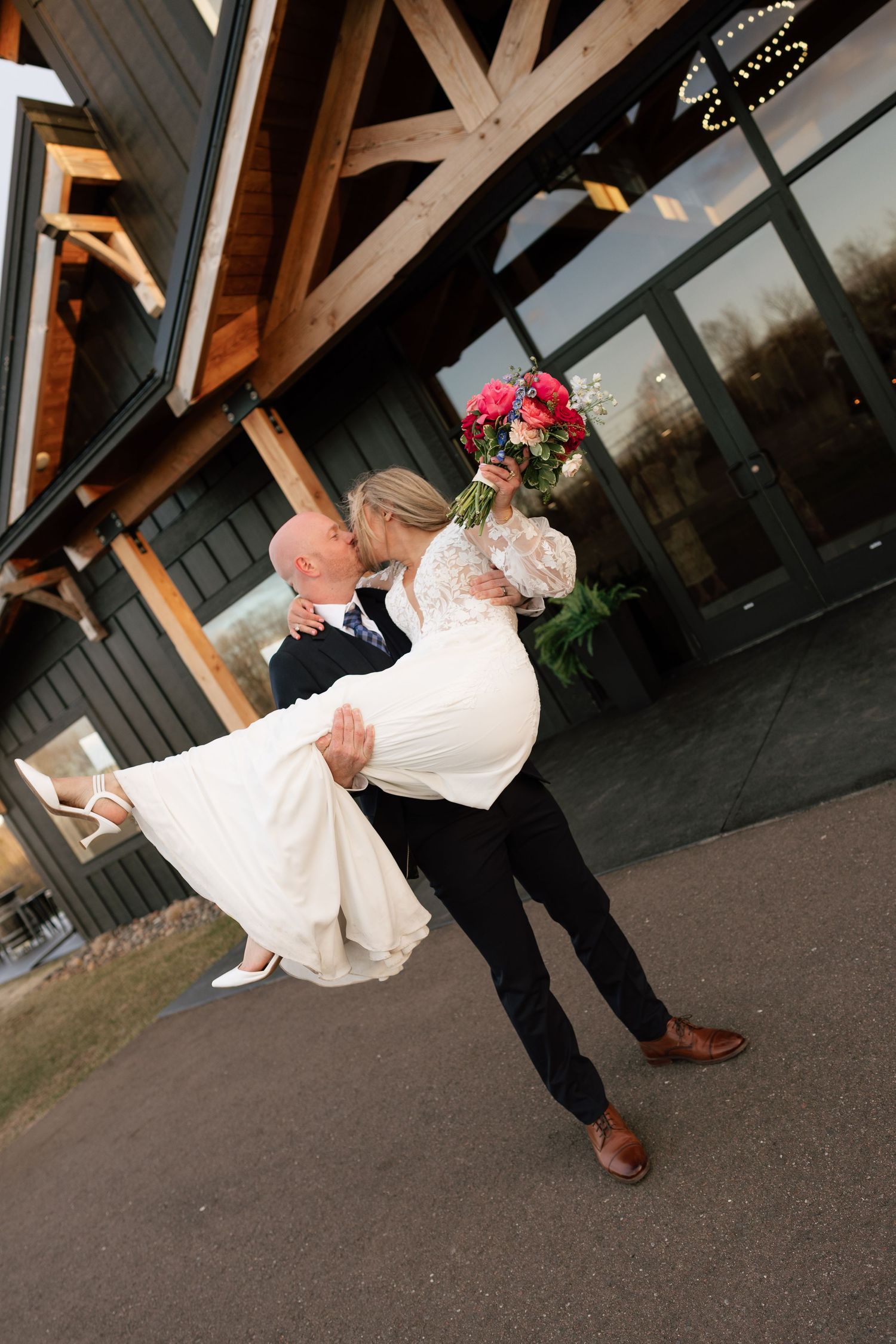 Groom lifts bride in white lace dress while they kiss, holding pink bouquet outside rustic venue.