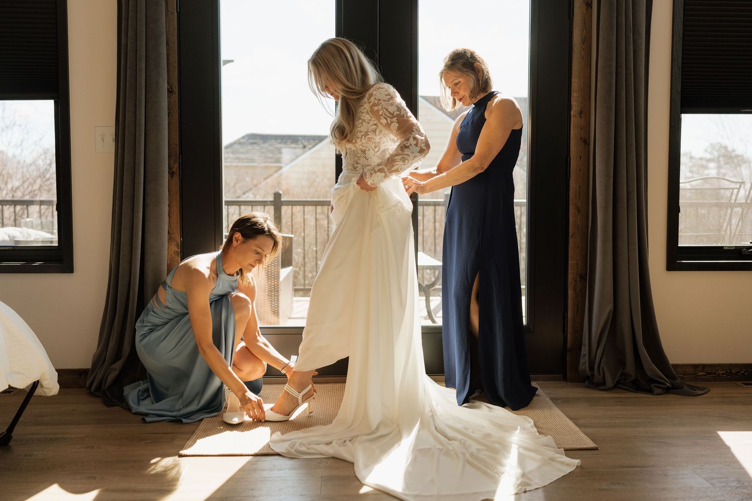 Bride getting ready as bridesmaids help with shoes and dress in sunlit room.