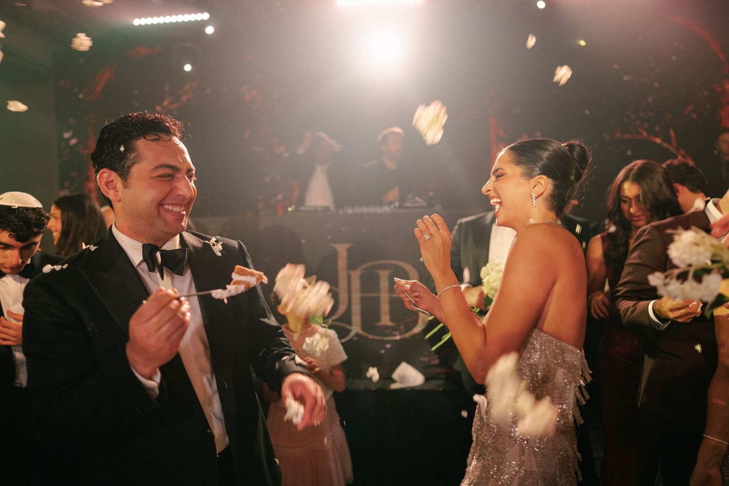 Smiling groom in a black tuxedo and bride in a sparkling gown clap and celebrate as white petals fly through the air at a wedding reception.