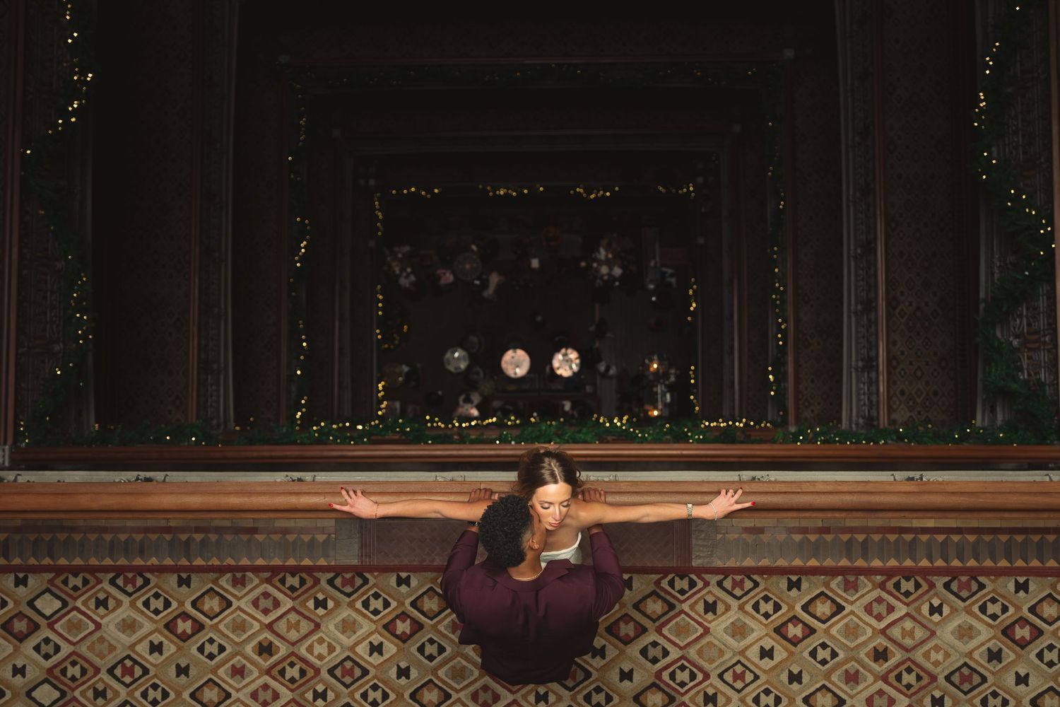 Woman in burgundy dress with arms outstretched facing ornate window with floral display and fairy lights.