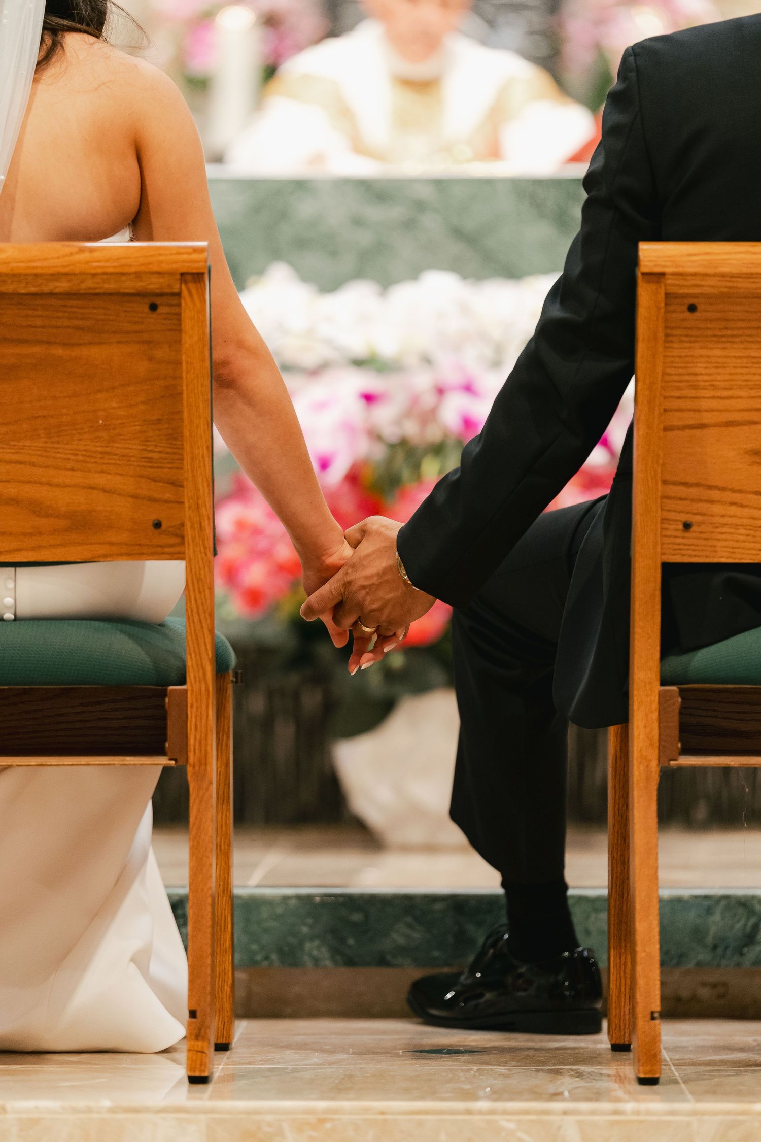 Bride and groom holding hands during wedding ceremony, seated in wooden chairs with floral altar and priest in background.