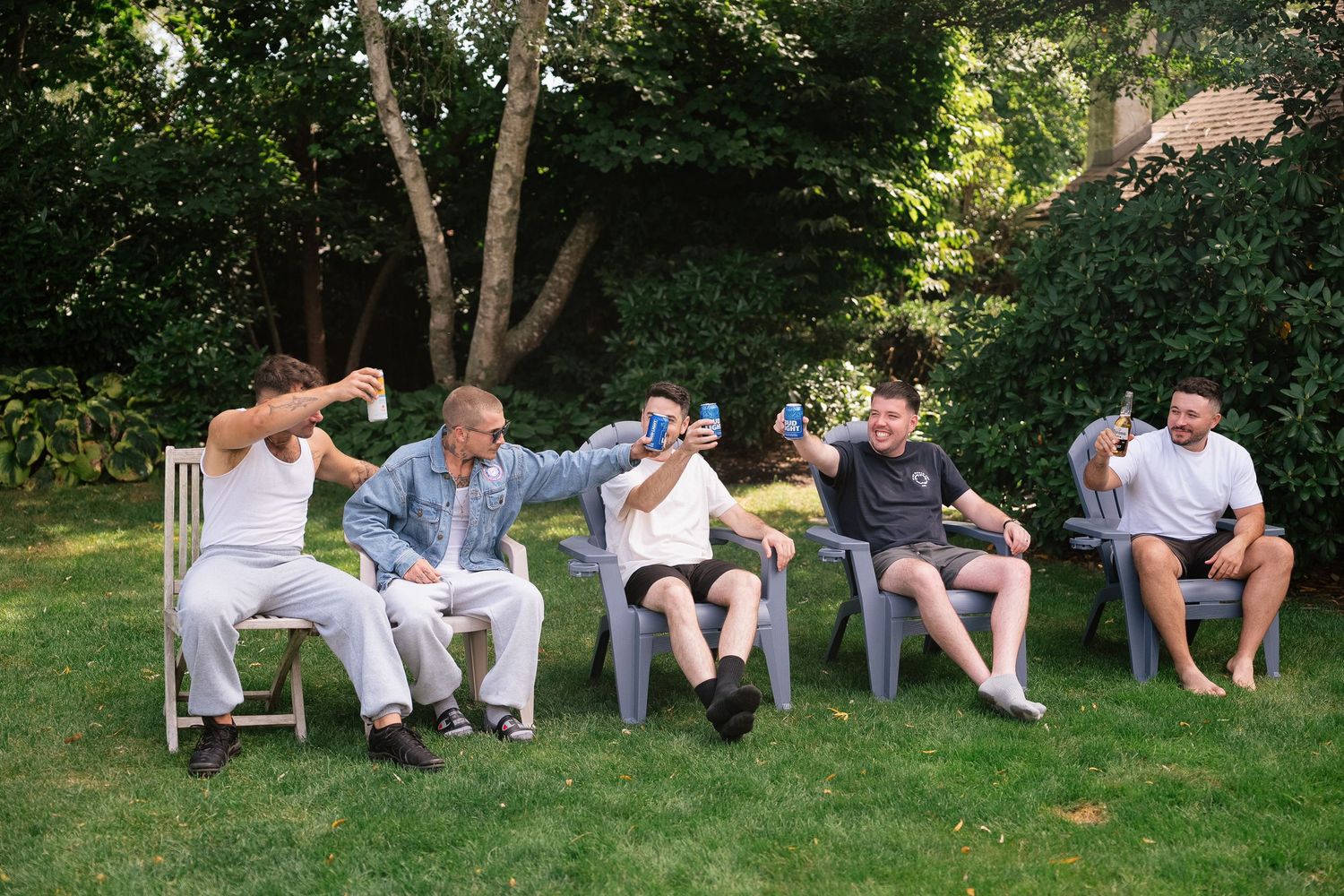 People sit casually on white chairs arranged in a row outdoors beneath green trees on a sunny day.