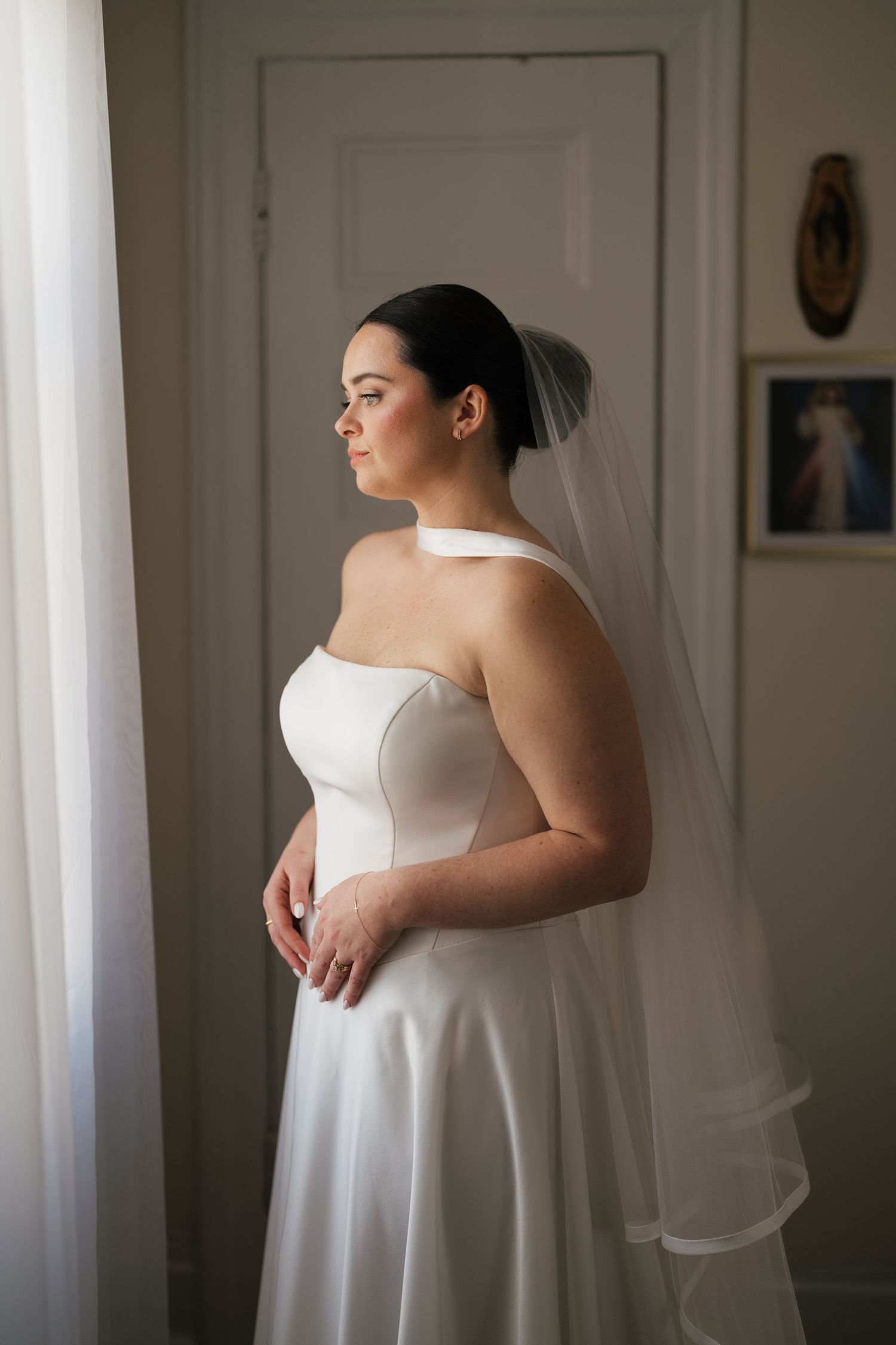 Bride in white strapless gown and veil stands by window, gazing thoughtfully in elegant room.