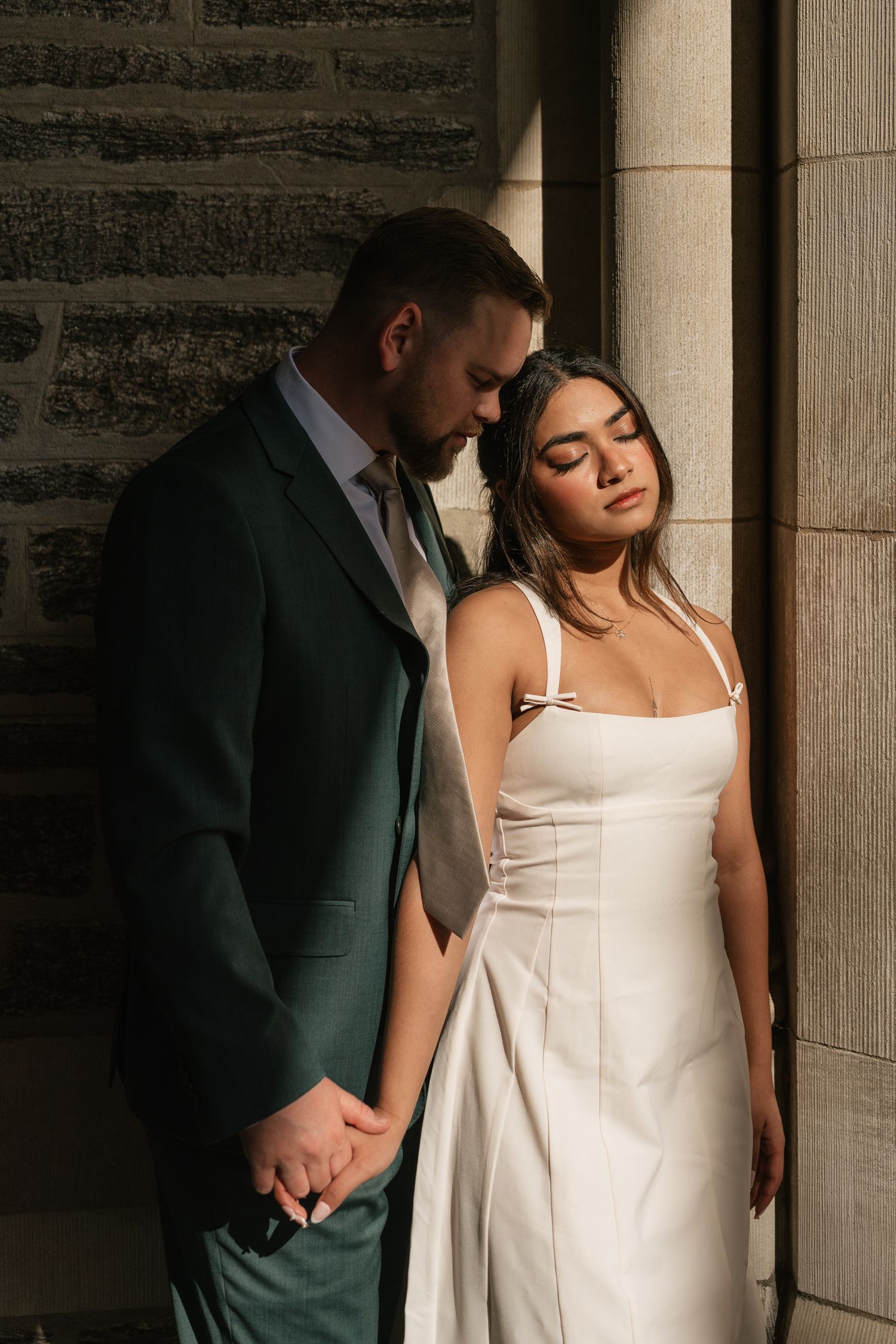 Bride in white gown and groom in dark green suit hold hands against stone columns in warm sunlight.