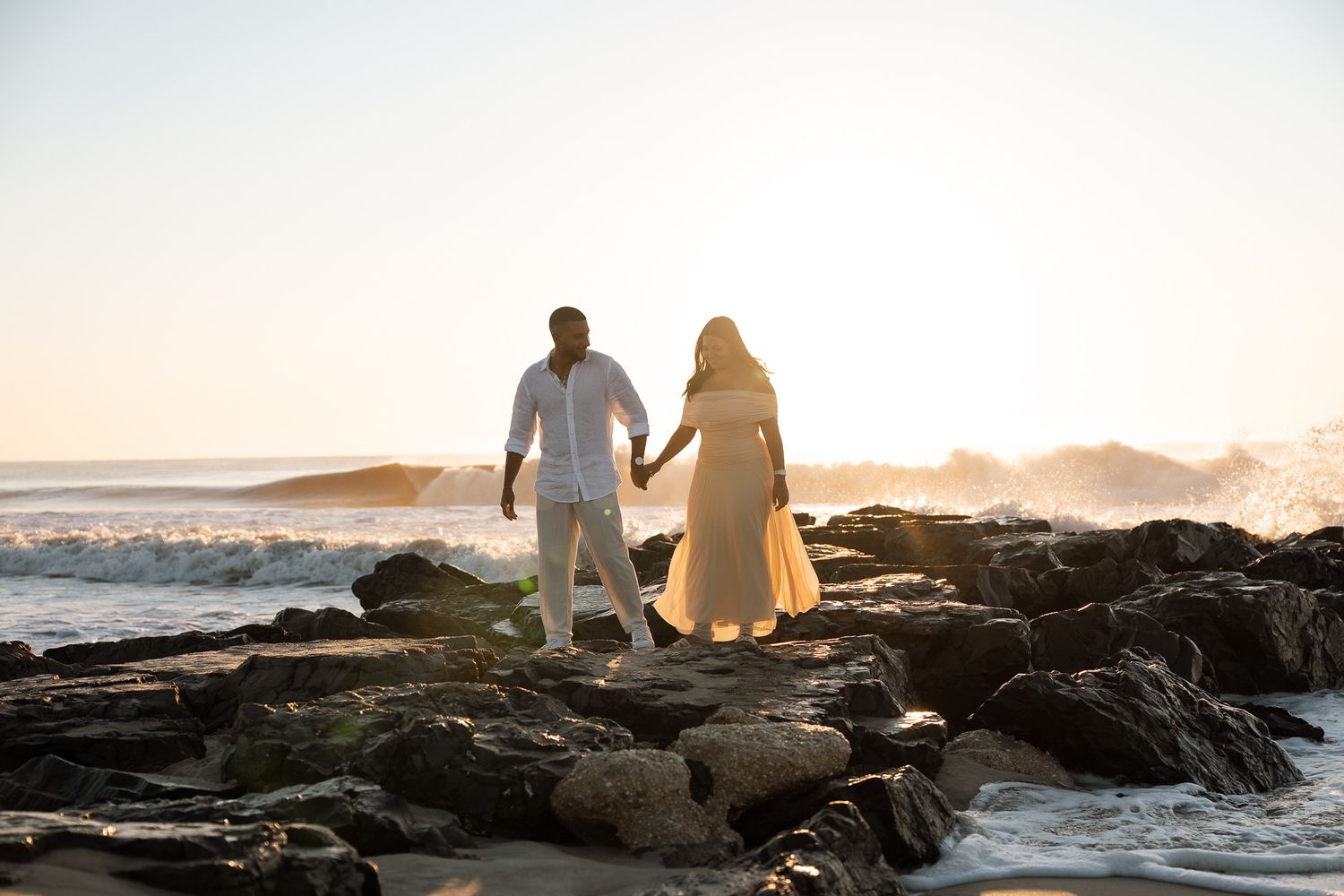 Couple walking along rocky coastline at sunset with ocean waves crashing in background.
