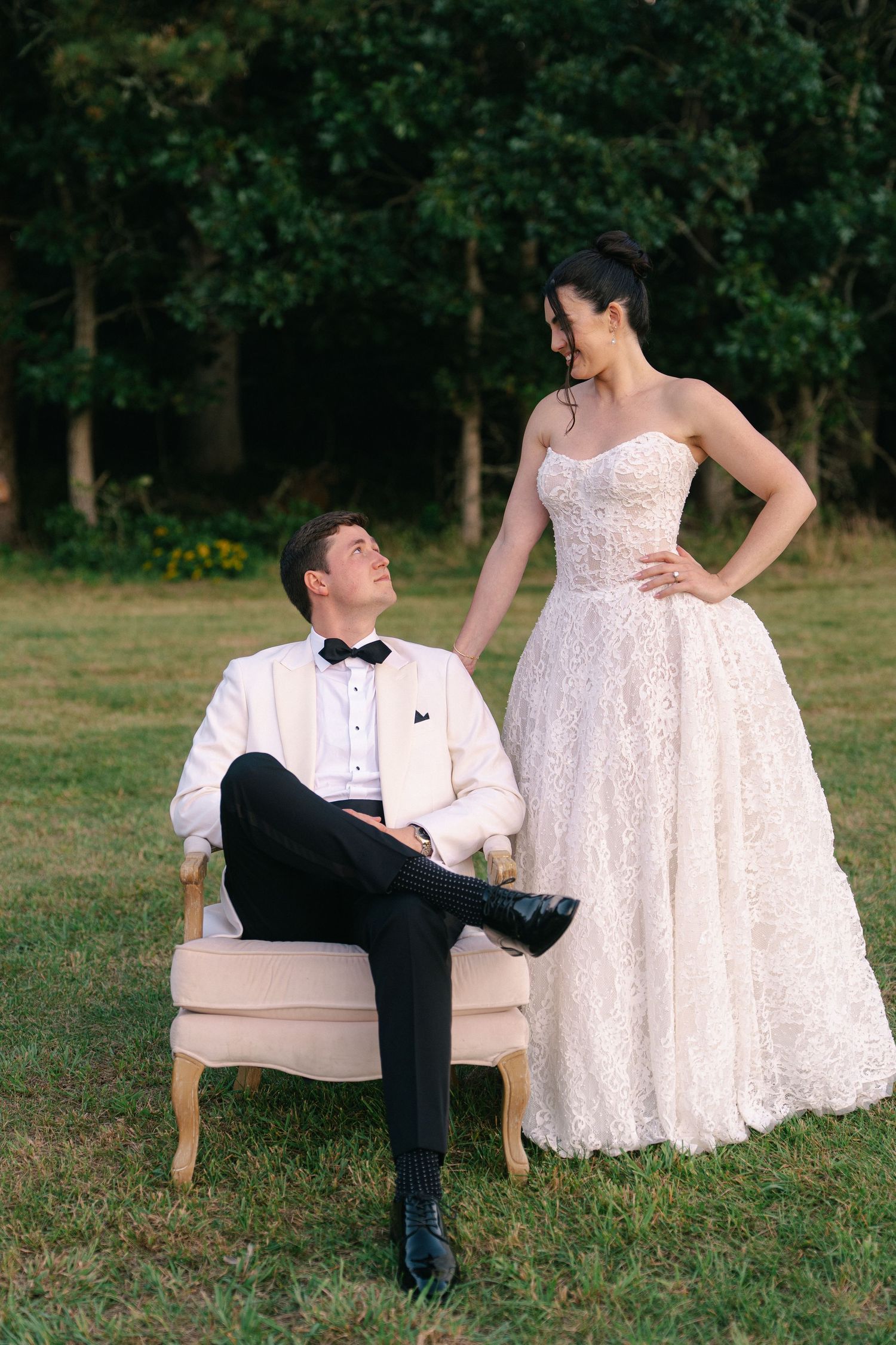 Bride in a lace strapless wedding gown stands beside a seated groom in a white tuxedo jacket and bow tie on a grassy lawn.