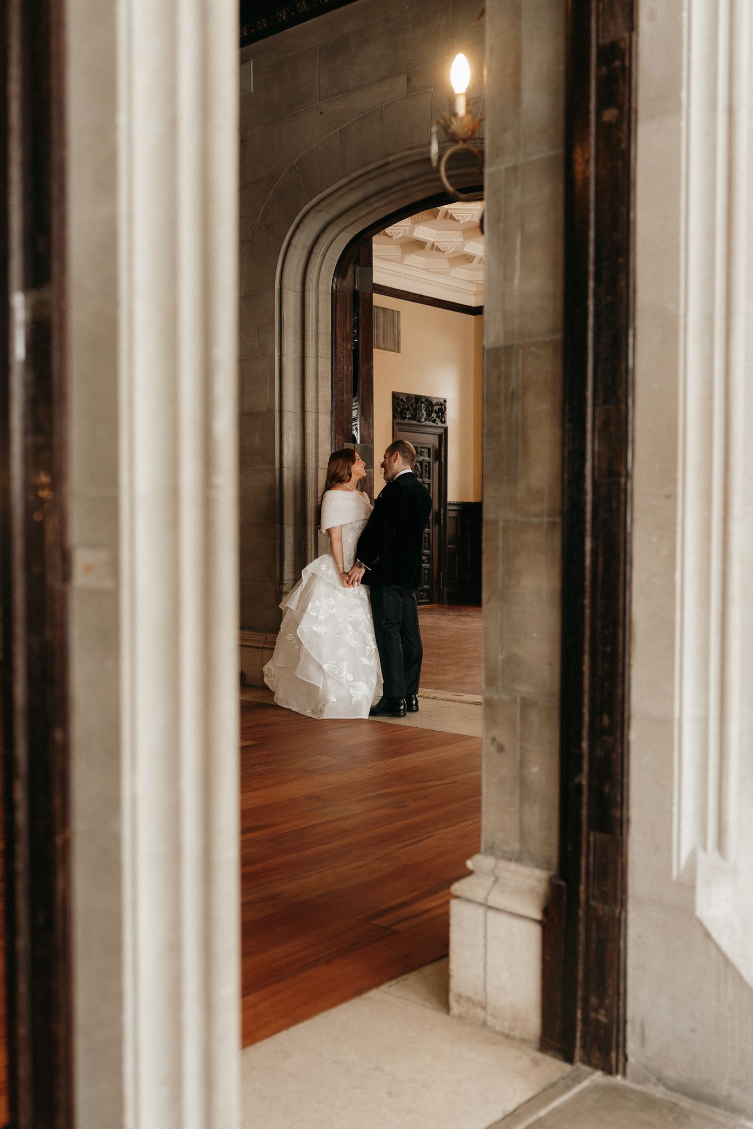A bride and groom share a romantic moment framed by an elegant stone archway in a grand historic venue.