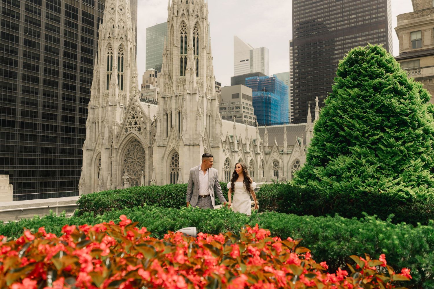 Couple walking hand in hand in front of St. Patrick's Cathedral in NYC, surrounded by lush greenery and red flowers.