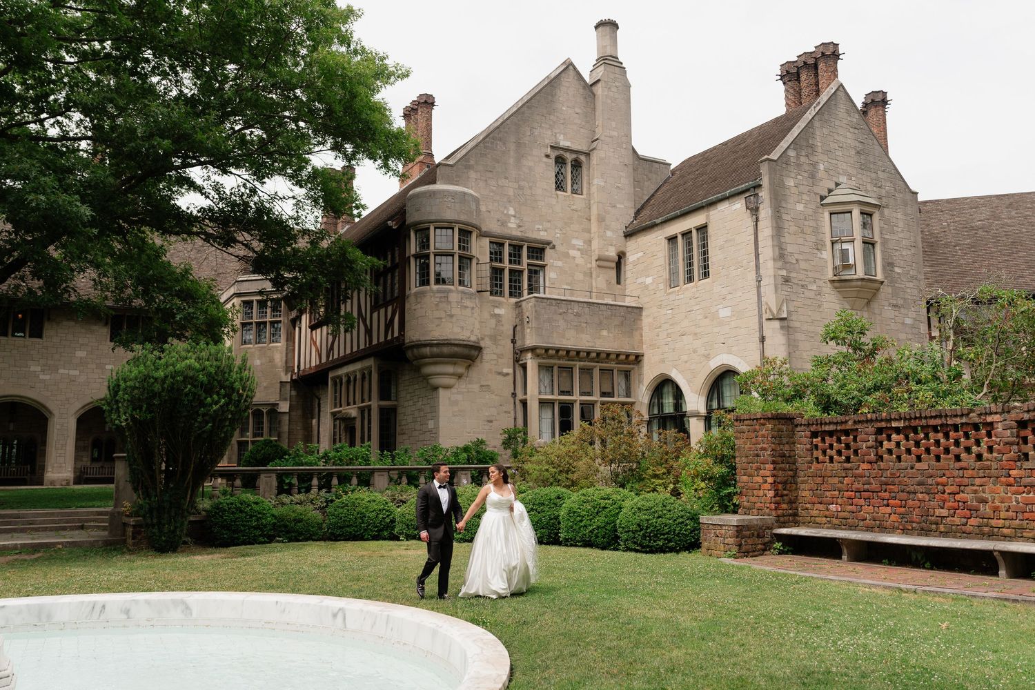 Bride and groom walking near fountain outside elegant stone manor wedding venue surrounded by lush gardens.