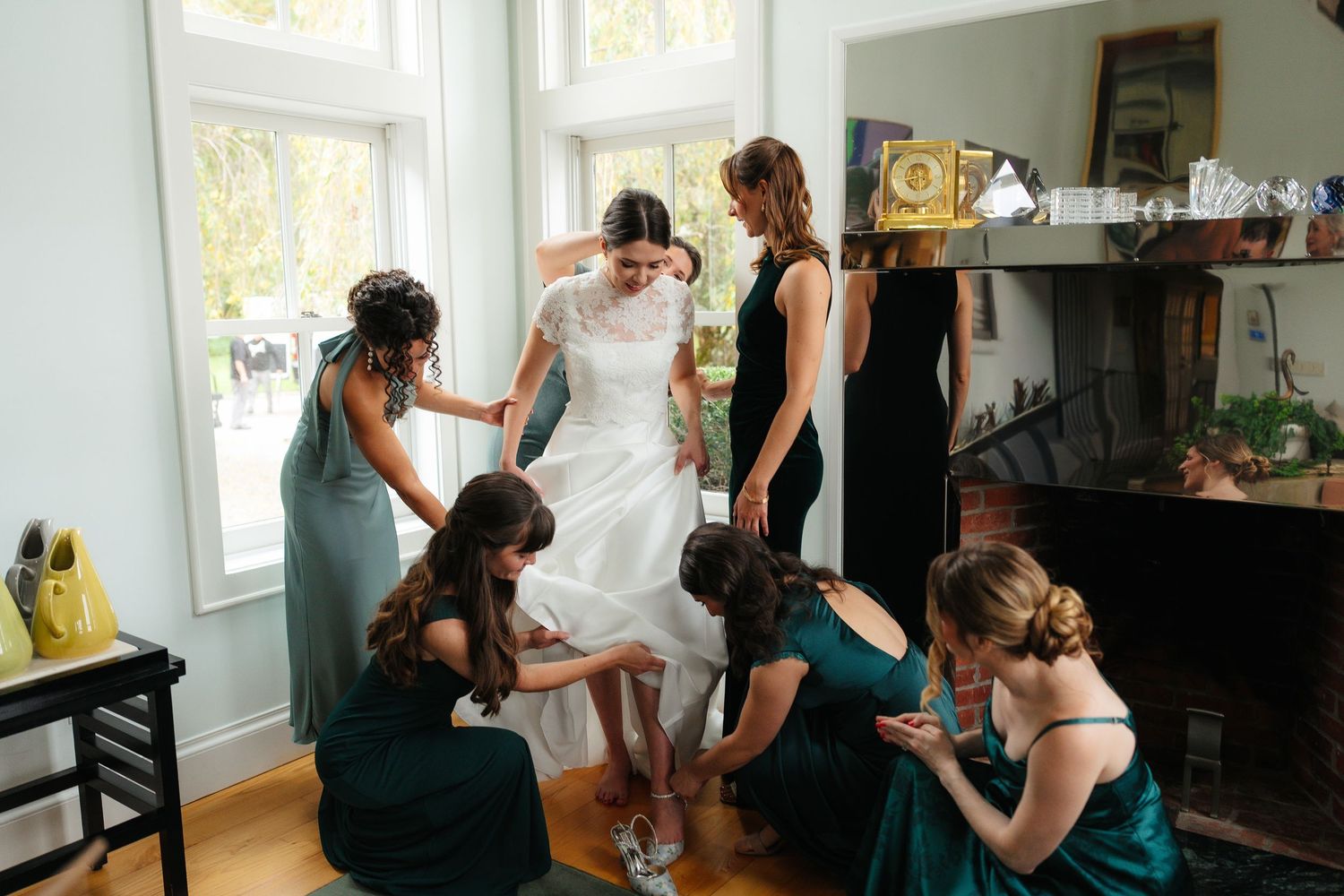 Bridesmaids in green dresses helping bride with shoes before wedding ceremony in elegant room.